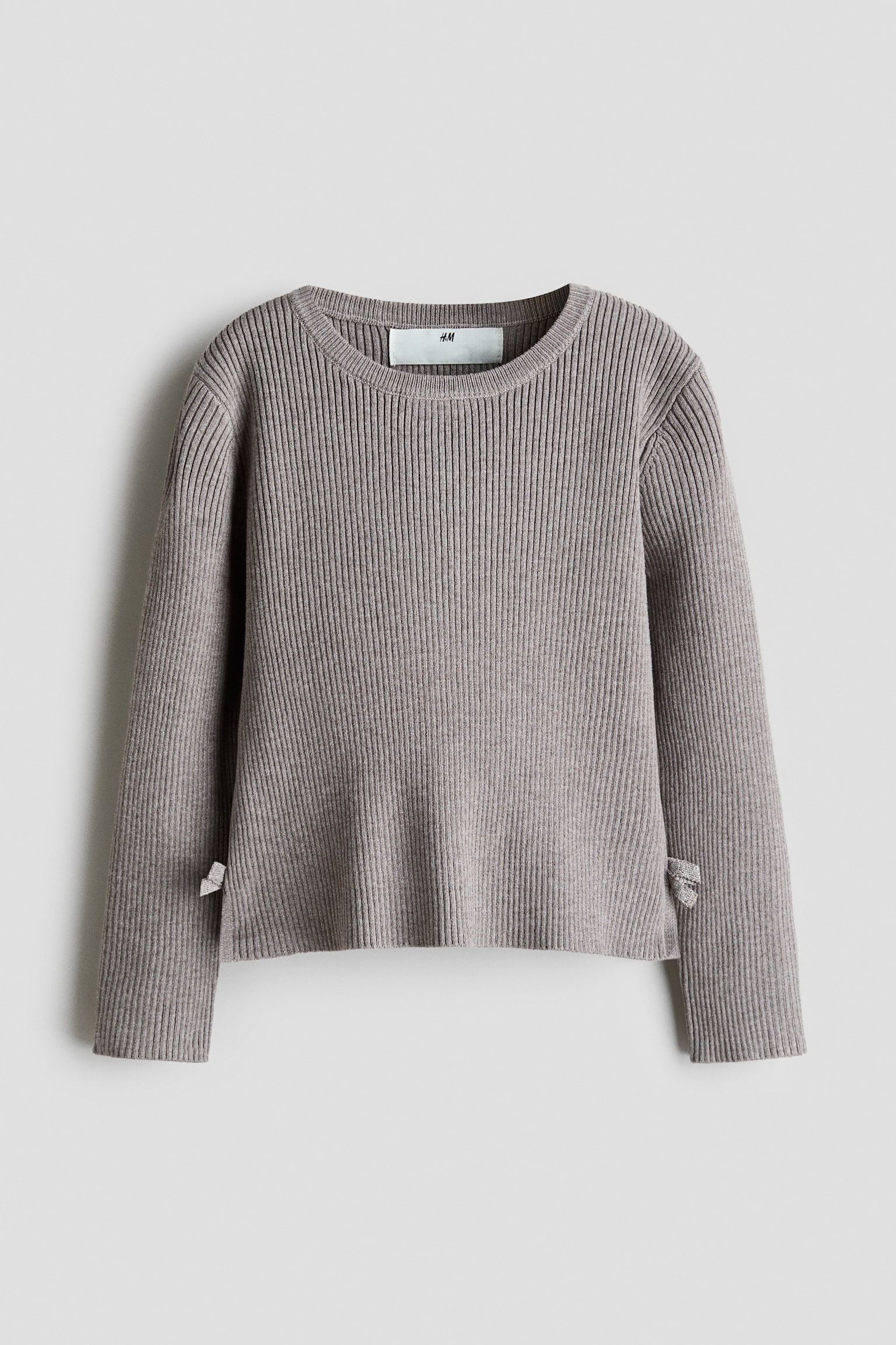 A dense rib-knit jumper in a muted taupe colour is shown from the front, featuring a round, rib-trimmed neckline, long sleeves, and a straight hem. The jumper is detailed with small side splits at the hemline that feature tiny, matching fabric bows.