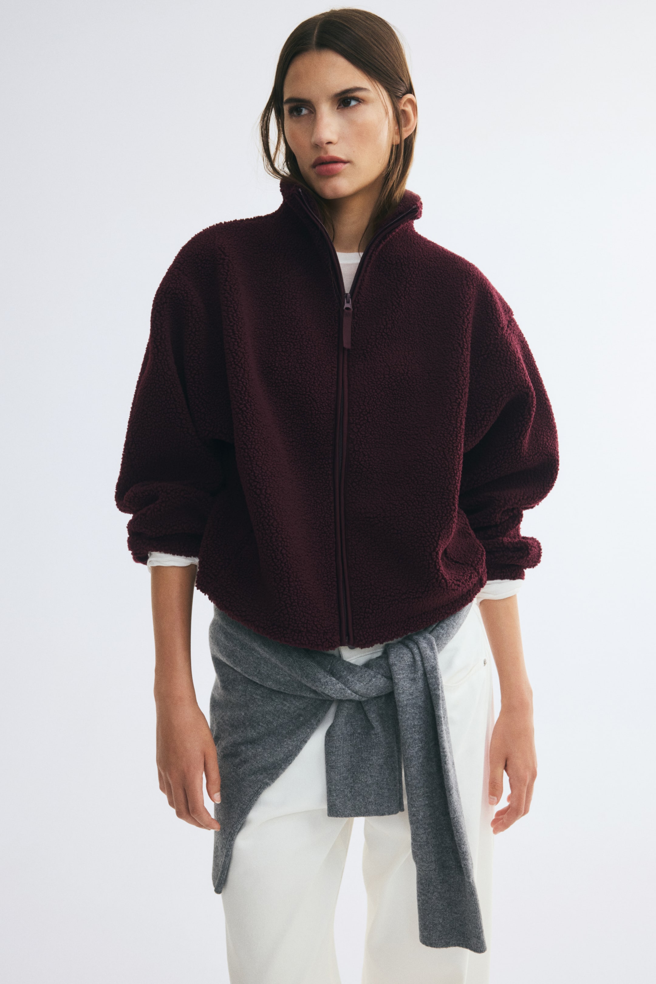 View larger image: A burgundy teddy fleece jacket featuring a stand collar, a front zipper, and dropped shoulders is worn by the model. The relaxed-fit jacket is layered over a white top and paired with white trousers and a grey sweater tied around the waist.