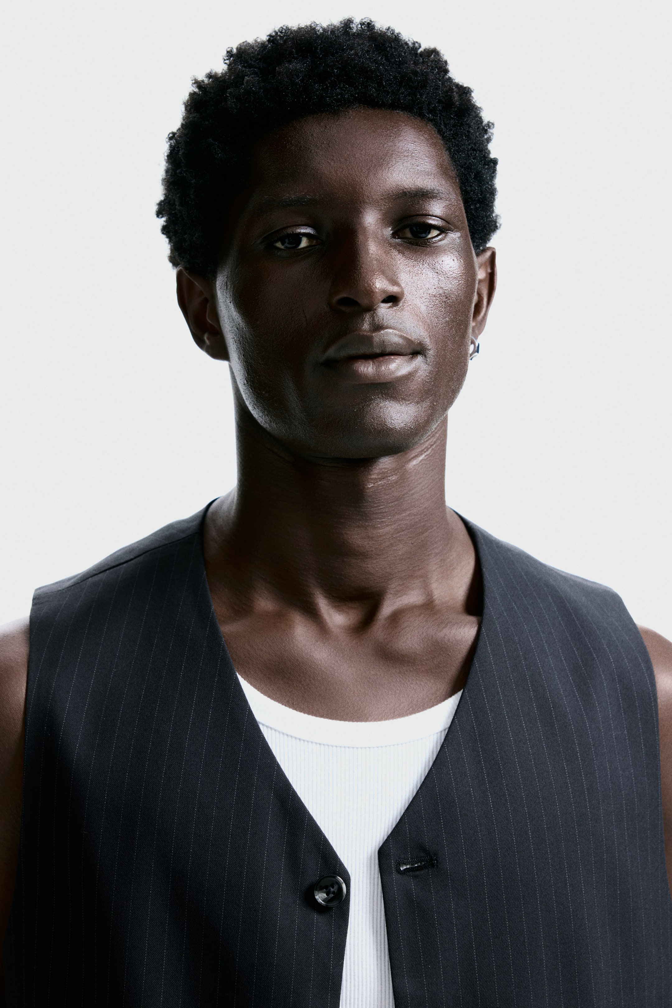 View larger image: A dark pinstripe vest is being shown from the front, layered over a white ribbed tank top. The vest is displaying a V-neckline and a single button closure at the lower front.