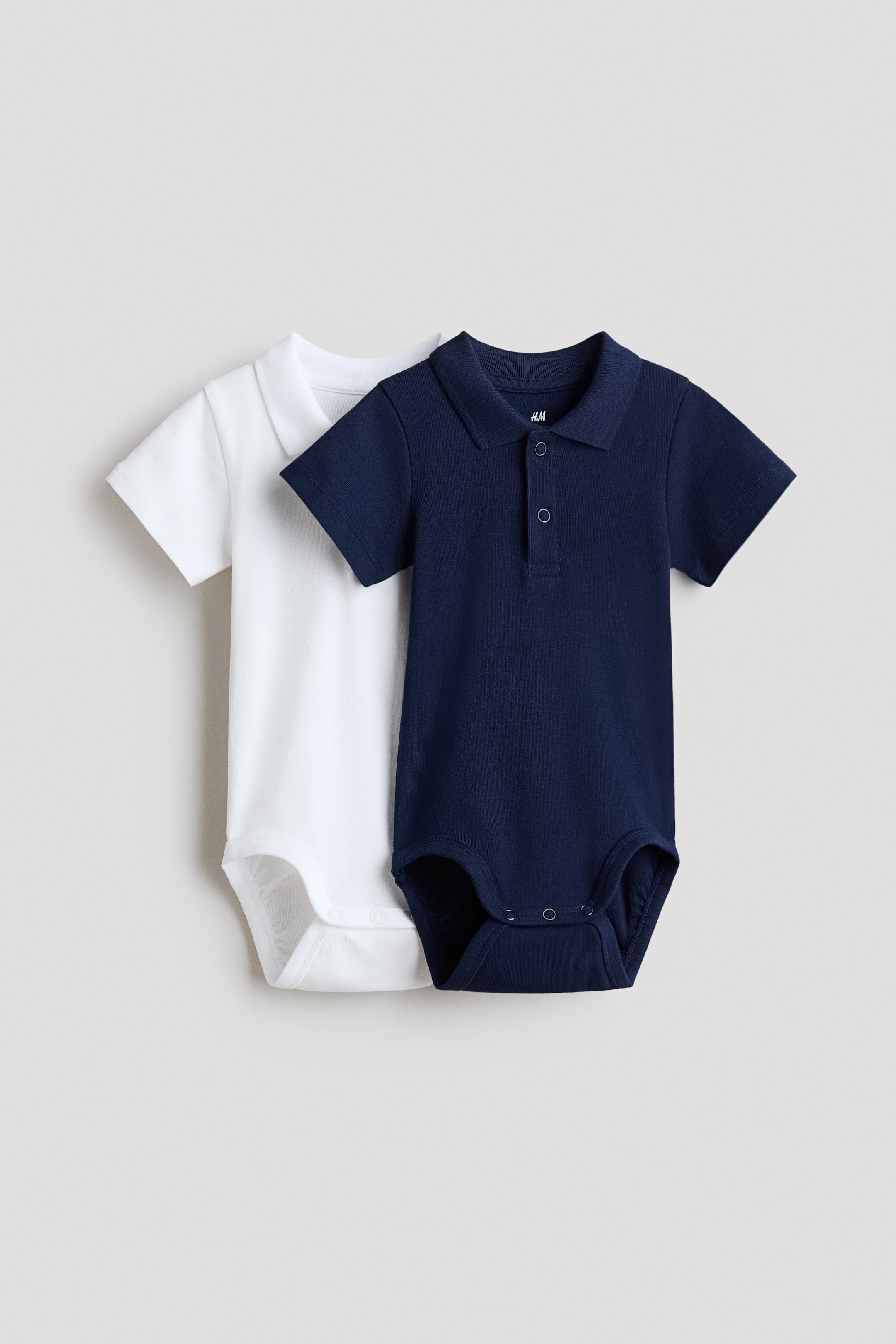 Grösseres Bild ansehen: Zwei Baby-Polo-Bodysuits werden präsentiert, wobei ein weißer teilweise hinter einem dunkelblauen liegt. Beide haben kurze Ärmel, klassische Kragen, eine Drei-Knopf-Leiste am Hals und Druckknöpfe im Schritt.