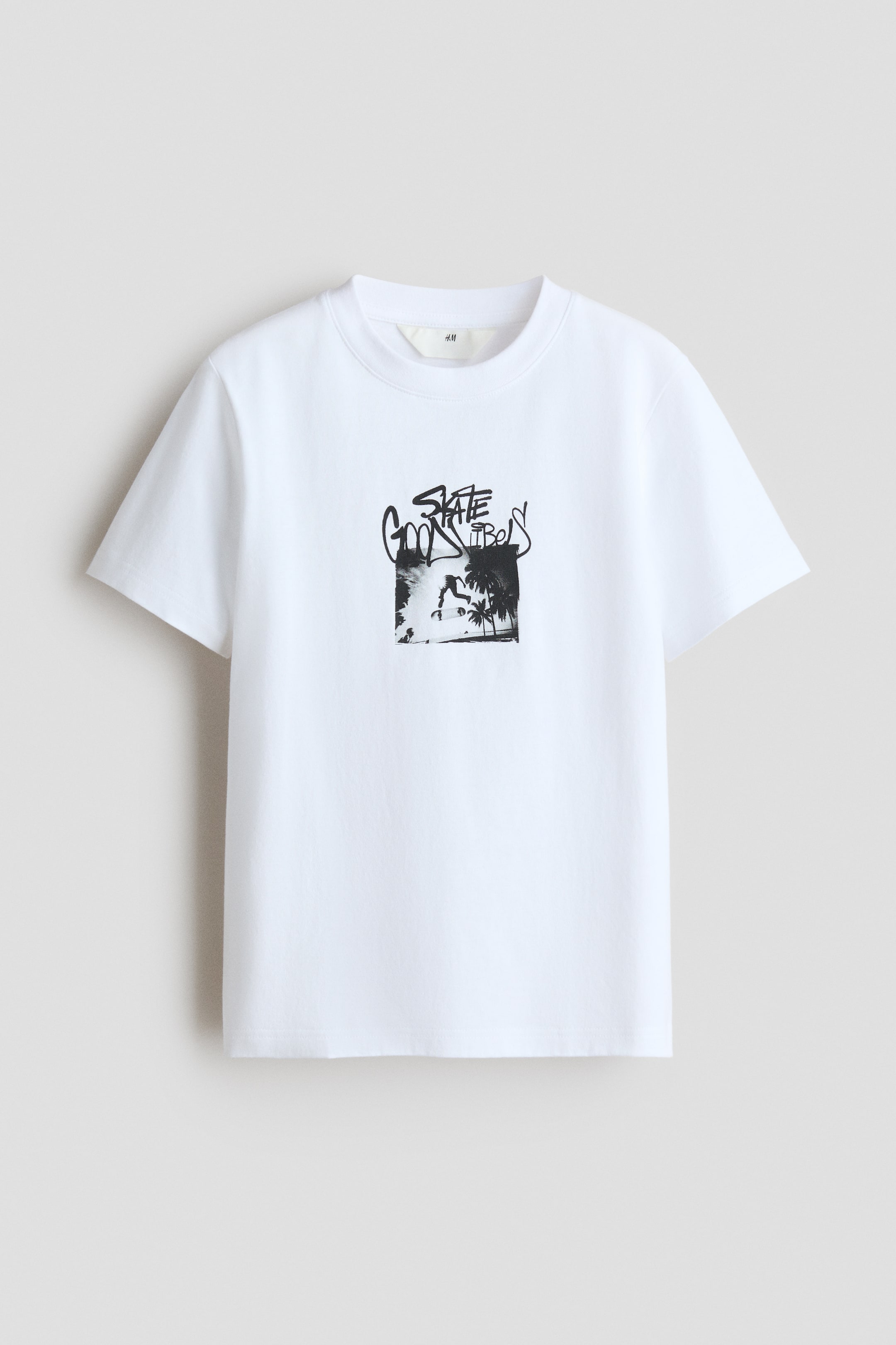 Agrandir l'image: Un t-shirt classique en jersey de coton blanc doux présente son devant, avec un imprimé graphique noir d'un skateur avec des palmiers, surmonté d'un texte de style graffiti indiquant "SKATE GOOD VIBES". Il est conçu avec des bords côtelés autour de l'encolure.