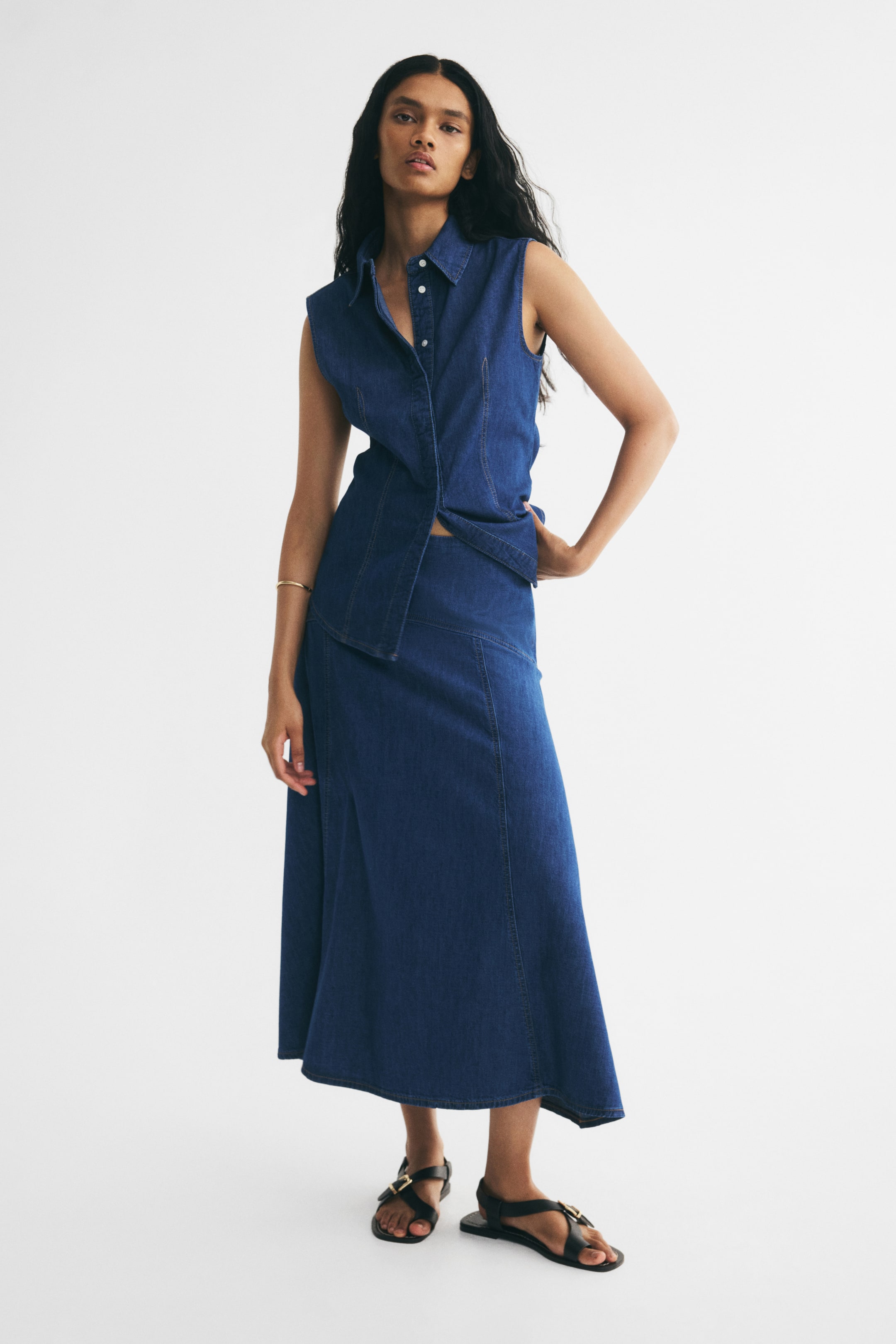 View larger image: A calf-length, A-line skirt in blue cotton denim is worn by the model, featuring a high waist, a curved seam across the hips, and vertical shaping seams down the front. The skirt is styled with a matching sleeveless denim top and black flat sandals.