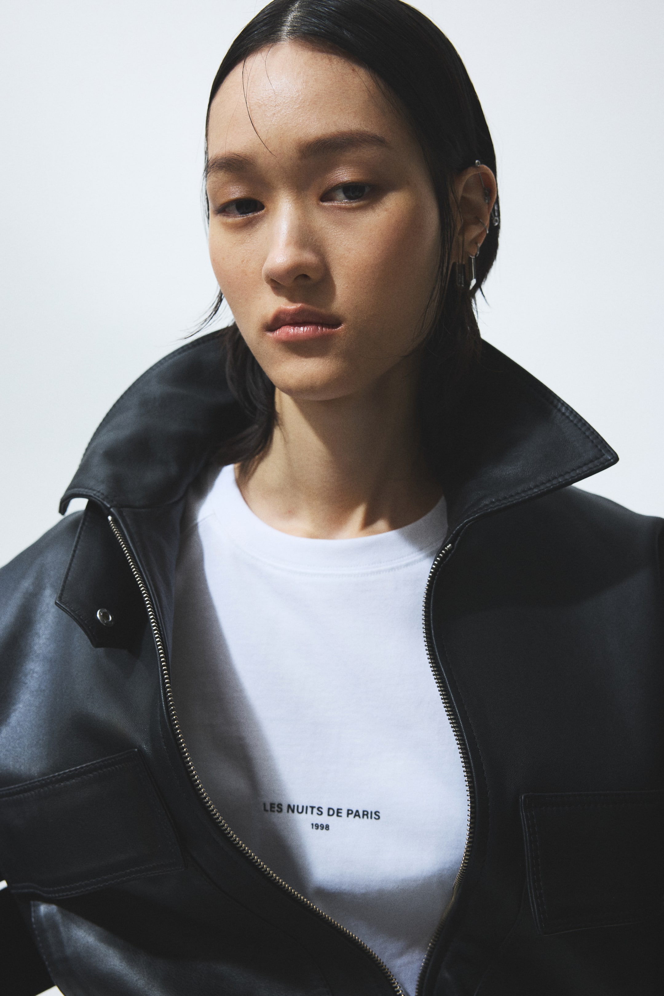 View larger image: A black jacket is worn open, slightly angled to the right, revealing a white crew-neck T-shirt underneath. The jacket features a wide collar, a visible silver zipper, a snap button, and a flap pocket on the chest. The T-shirt displays "LES NUITS DE PARIS 1998" printed in black on the front.