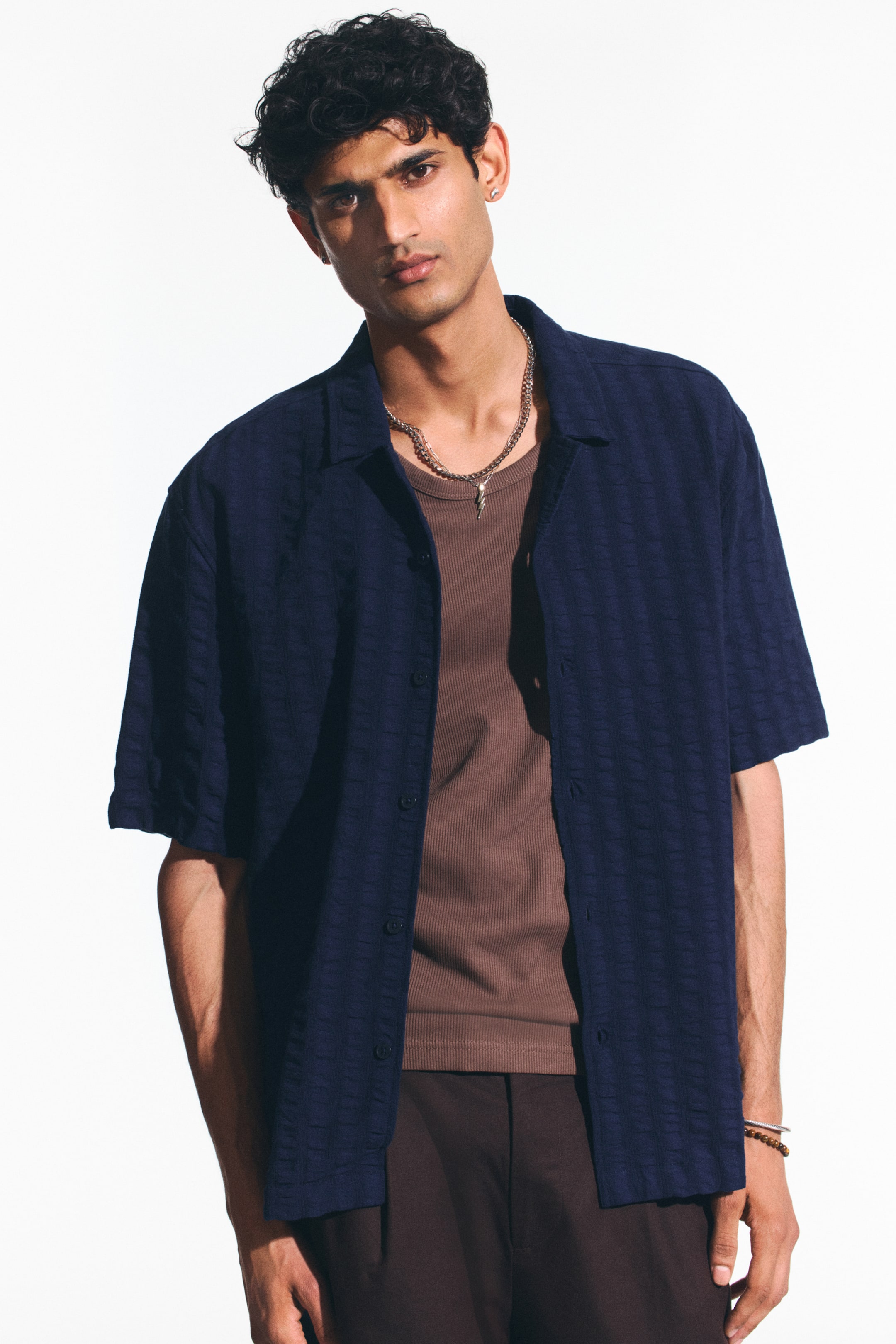 View larger image: A navy blue short-sleeved shirt is worn open over a brown ribbed t-shirt. The shirt showcases a subtle, raised geometric pattern throughout, a spread collar, and dark buttons down the front.