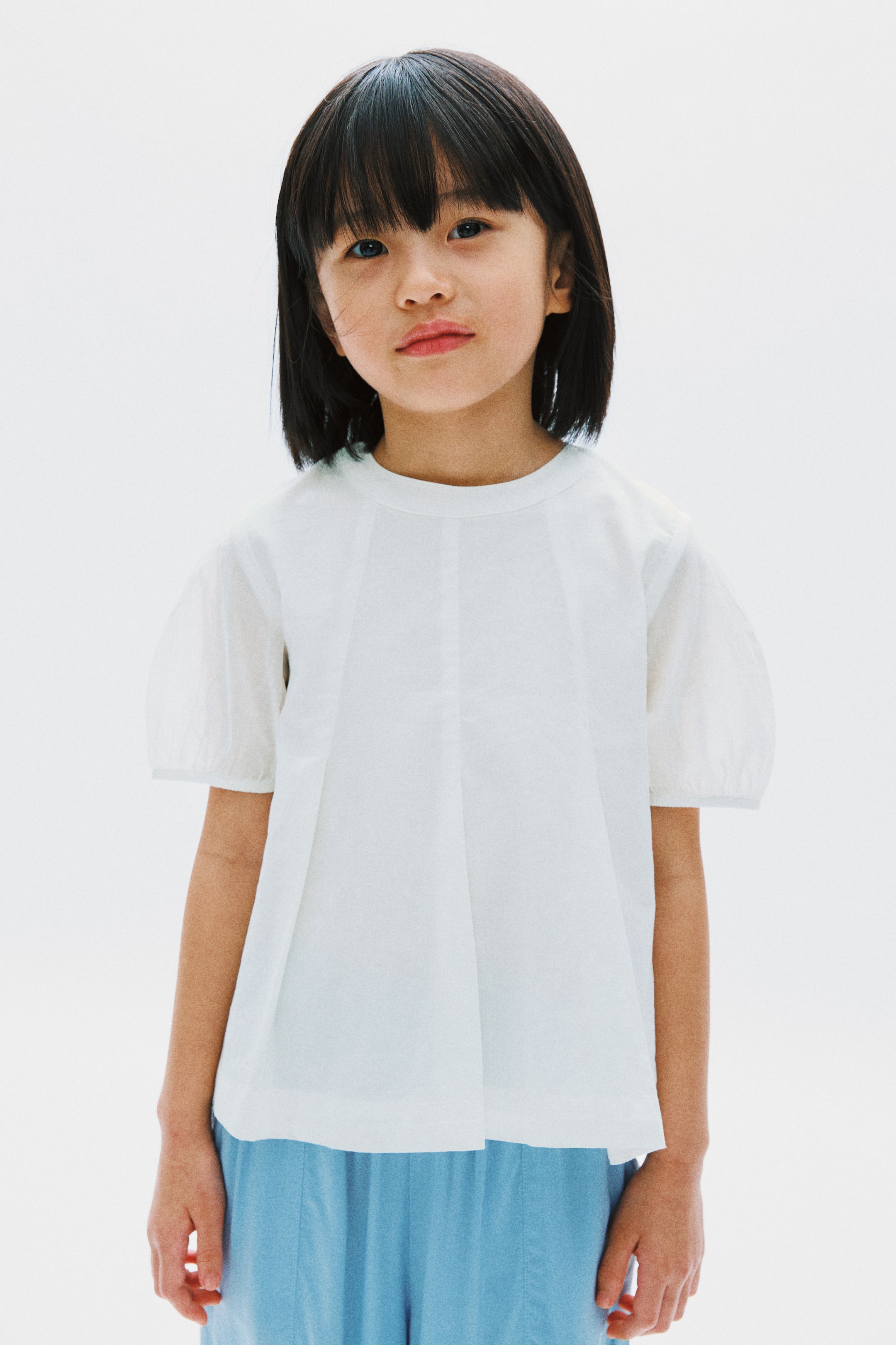 View larger image: The white short-sleeved top features a round neckline and puffed sleeves with gathered hems. It showcases multiple vertical design lines on the front and offers a loose, structured fit when facing forward.