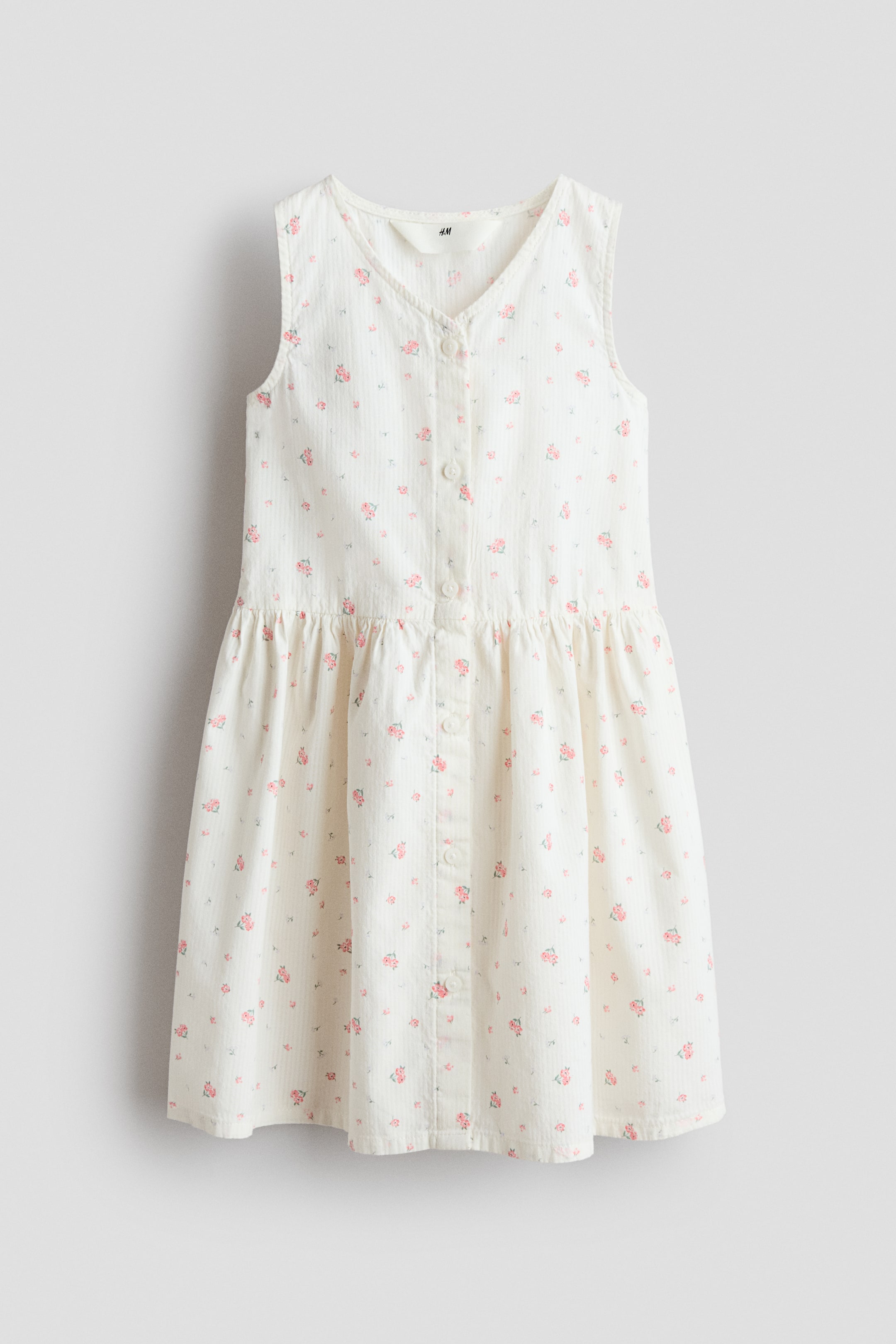 Größeres Bild ansehen: Ein ärmelloses Kleid aus gemustertem Baumwoll-Seersucker präsentiert seine Vorderseite mit einem V-Ausschnitt, einer Knopfleiste vorne, einer gerafften Naht an der Taille und einem ausgestellten Rock. Das Kleid zeigt kleine rosa Blumenmuster auf weißem Hintergrund.