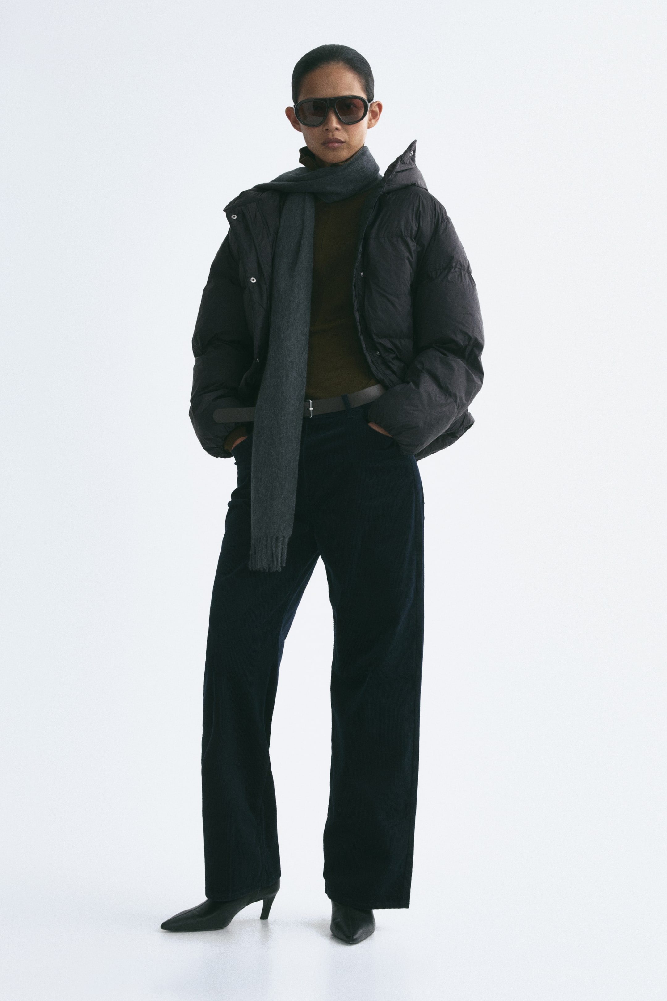 View larger image: The model is wearing a black hooded puffer jacket left open to reveal a layered outfit underneath. The jacket is styled with a dark brown high-neck top, a gray fringed scarf, and dark wide-leg trousers.