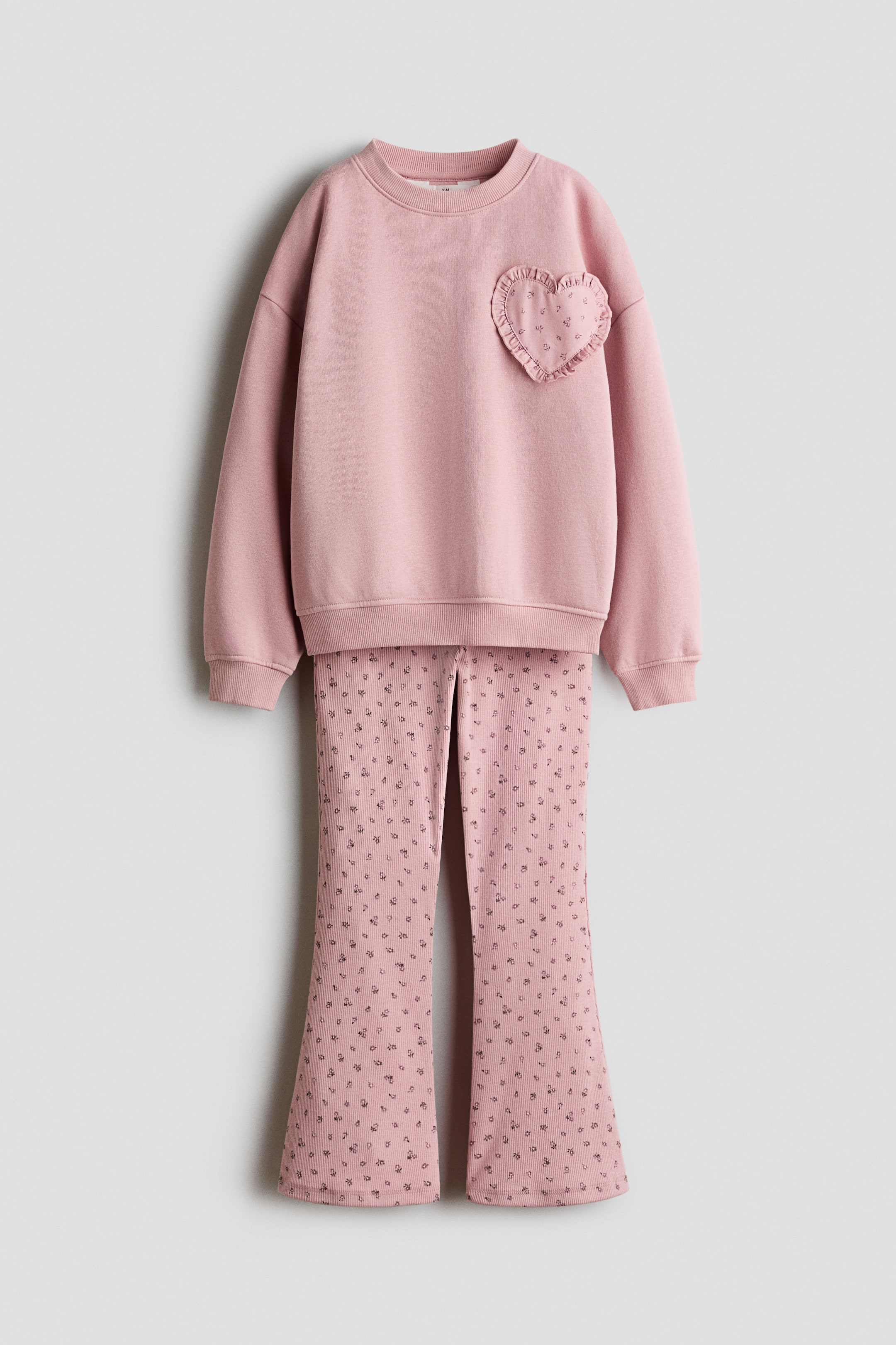 View larger image: A pink two-piece outfit showcases a sweatshirt and flared pants, both facing forward. The crewneck sweatshirt features long sleeves with ribbed cuffs and a heart-shaped patch with ruffled edges and a subtle floral pattern on the left chest. The matching flared pants display a ribbed texture with a scattered dark floral pattern.