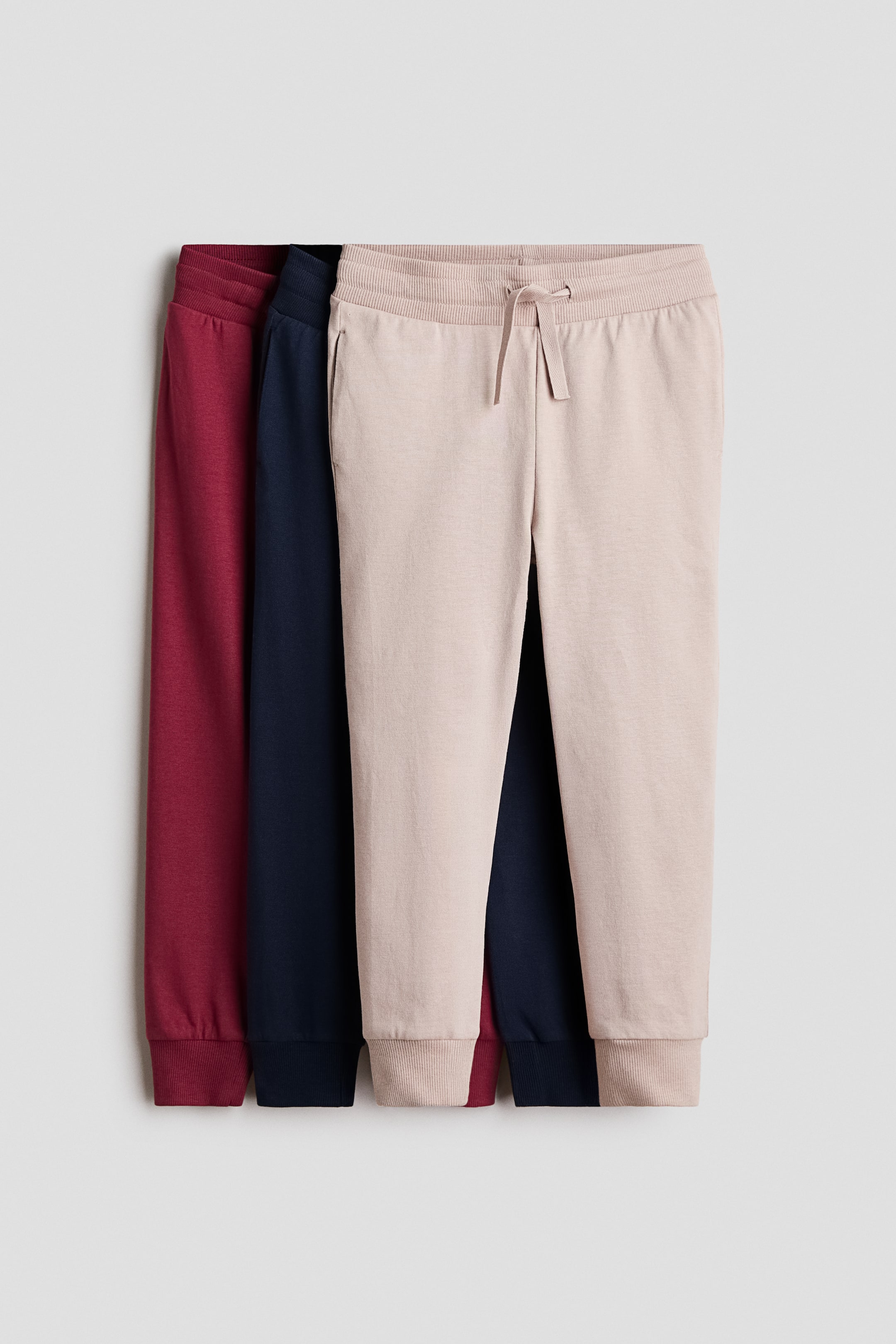 View larger image: A three-pack of cotton jersey joggers in burgundy, navy blue, and light beige is hanging in a layered, front-facing arrangement. The front pair is displaying an elasticated waistband with a drawstring, discreet side pockets, and ribbed cuffs.