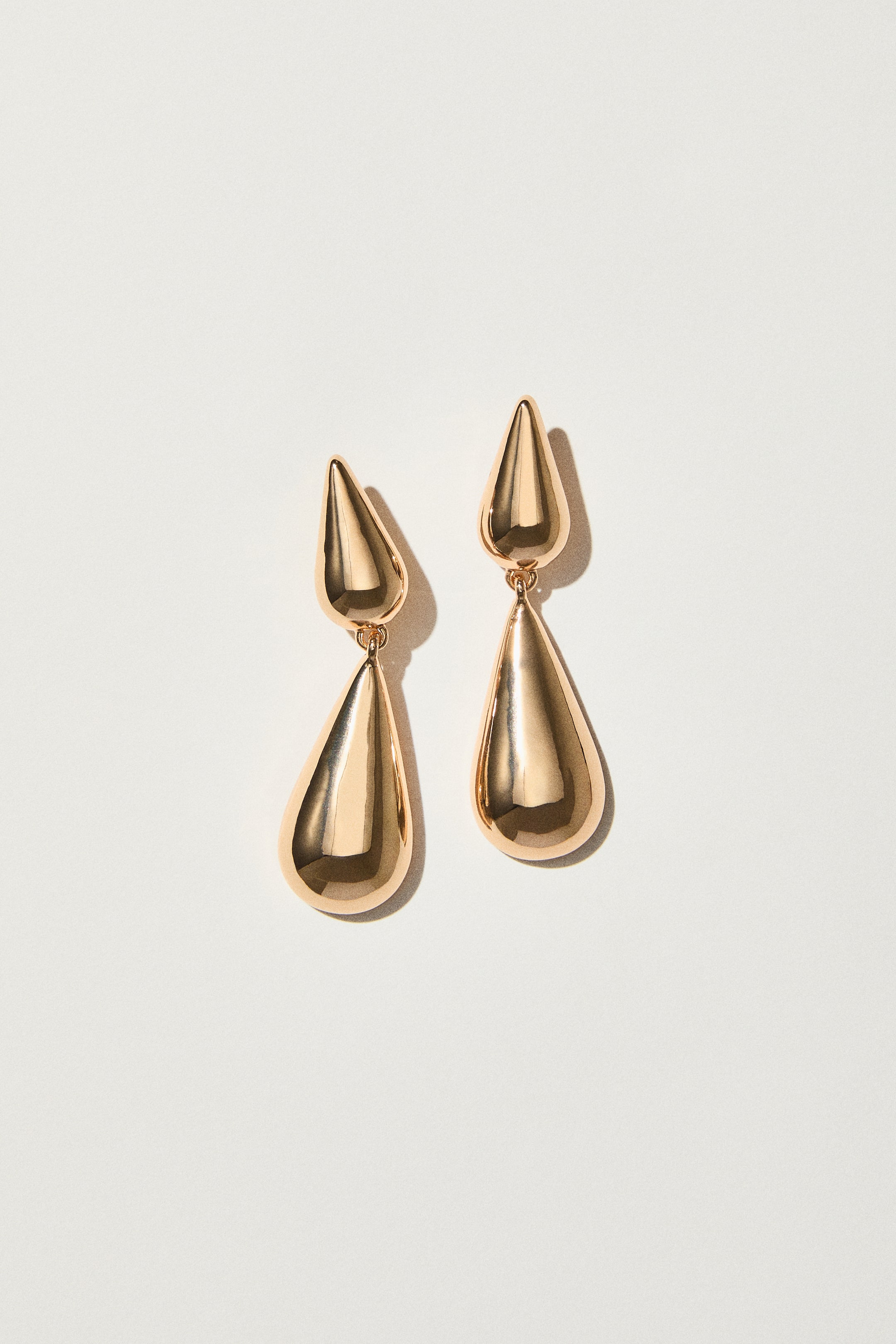 Ver imagen más grande: Dos pendientes de metal con tono dorado presentan un diseño frontal, con cada pendiente mostrando dos elementos en forma de gota conectados. Una gota más pequeña e invertida forma la pieza superior, que se conecta a un colgante más grande con forma de gota estándar.