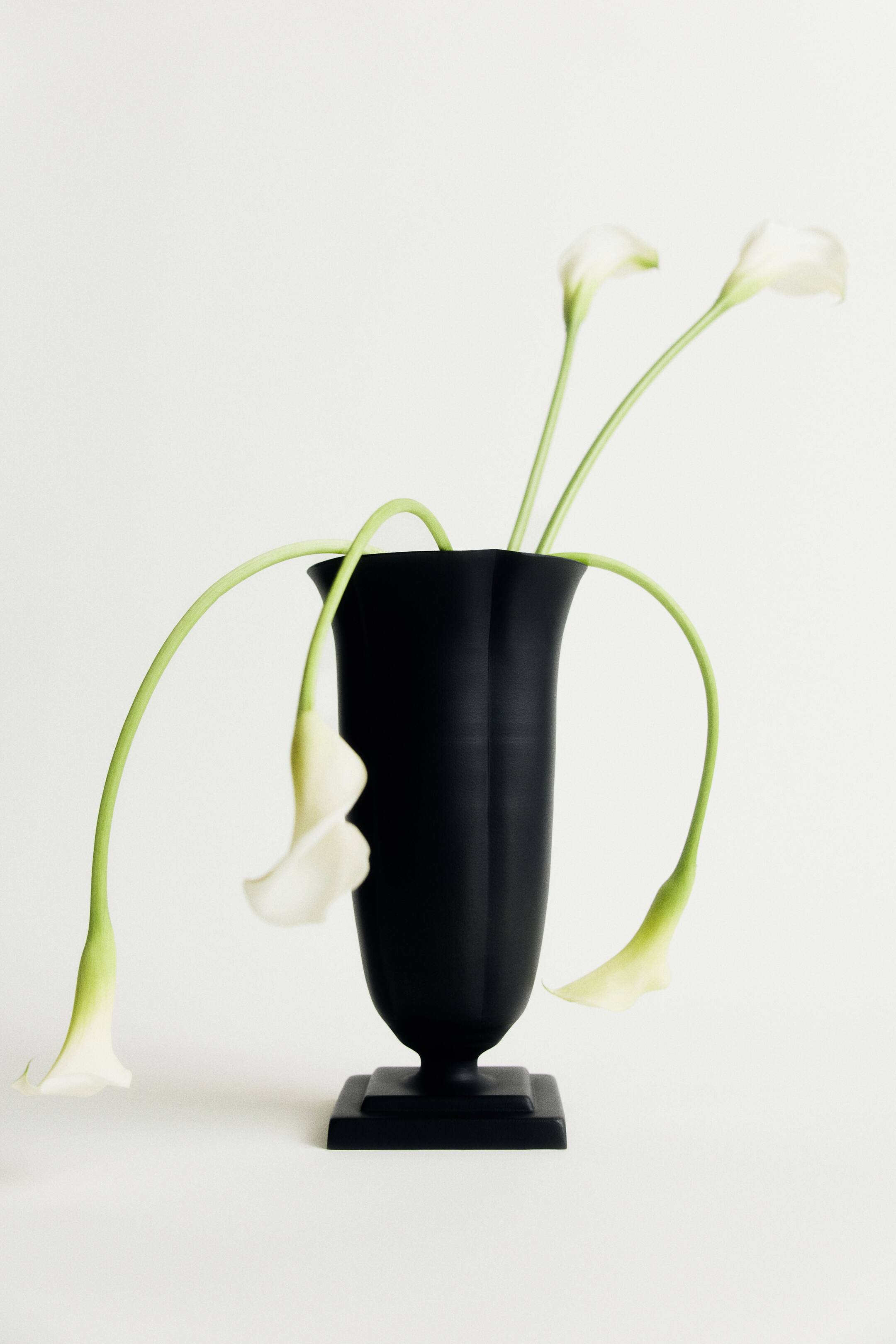Agrandir l'image: Un vase en métal noir mat, doté d’un bord cannelé et d’un pied sculptural carré à gradins, est présenté de face. Plusieurs arums blancs aux longues tiges vertes émergent de l’ouverture, certaines tiges retombant gracieusement le long des côtés du vase.