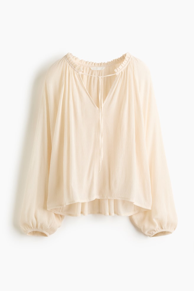 Una blusa de color crema muestra su parte frontal, con un escote fruncido en forma de V y detalles de lazo. Presenta mangas largas y voluminosas con puños elásticos y una textura arrugada en toda la prenda, ofreciendo un ajuste relajado y un dobladillo ligeramente curvado. - 1