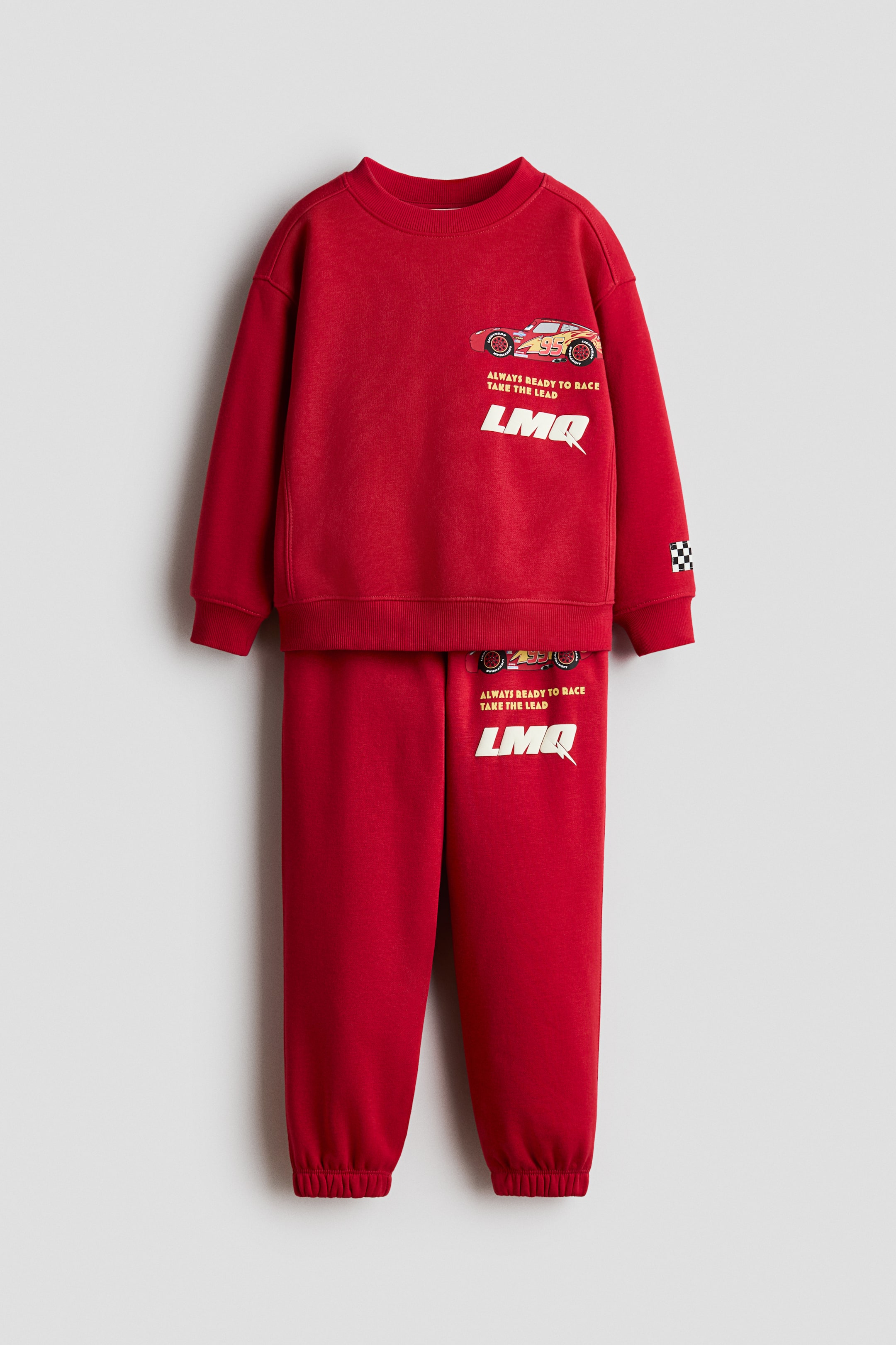 View larger image: A red tracksuit is being displayed front-facing, comprising a crew neck sweatshirt and matching joggers. The sweatshirt features an orange race car graphic with the number 95, "ALWAYS READY TO RACE TAKE THE LEAD" and "LMQ" lettering on the chest, a checkered flag patch on the left sleeve, and comes with ribbed cuffs and hem. The joggers showcase ribbed ankle cuffs and a coordinating race car graphic and lettering on the left leg.