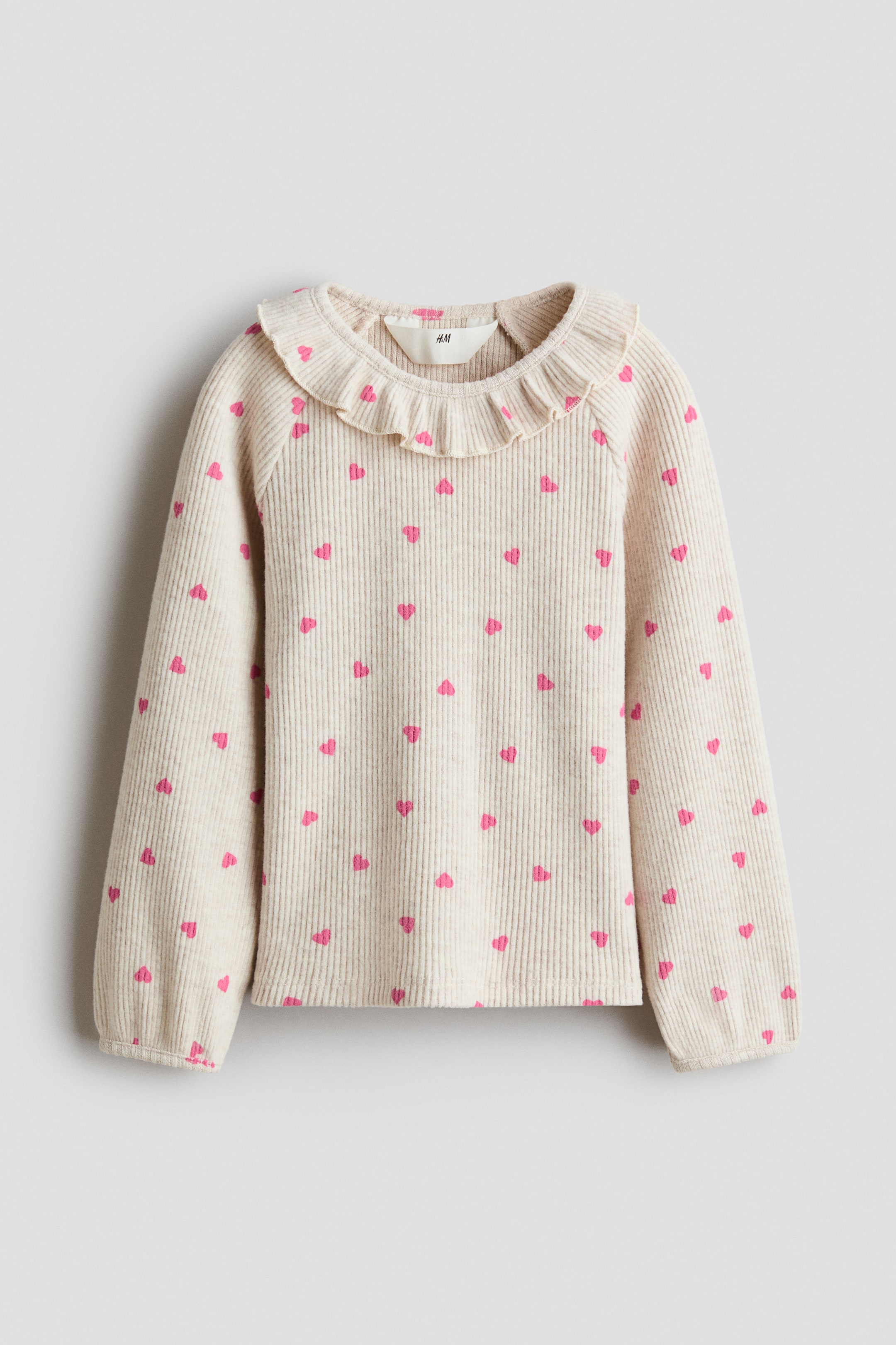 Agrandir l'image: Vue de face d’un haut en jersey de coton côtelé beige clair, présentant un motif intégral de petits cœurs roses. Le haut est doté d’un col rond à bordure volantée et de manches longues raglan se terminant par des poignets étroits bordés.