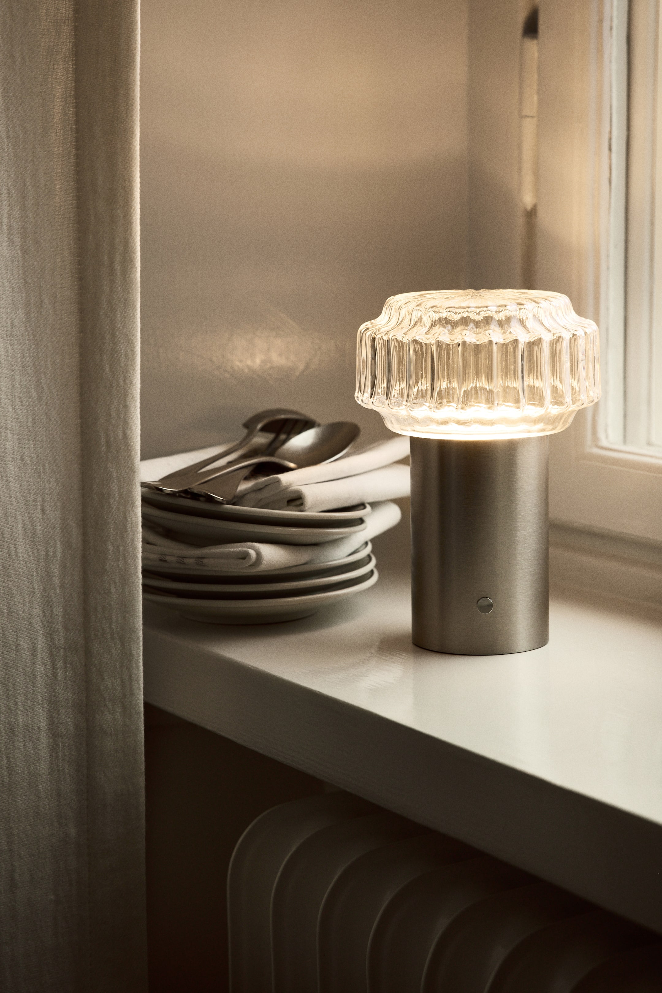 Agrandir l'image: Une lampe de table rechargeable est dotée d'une base cylindrique en métal et d'un abat-jour en verre transparent cannelé, qui diffuse la lumière. Un petit interrupteur rond marche/arrêt est visible à l'avant de la base.