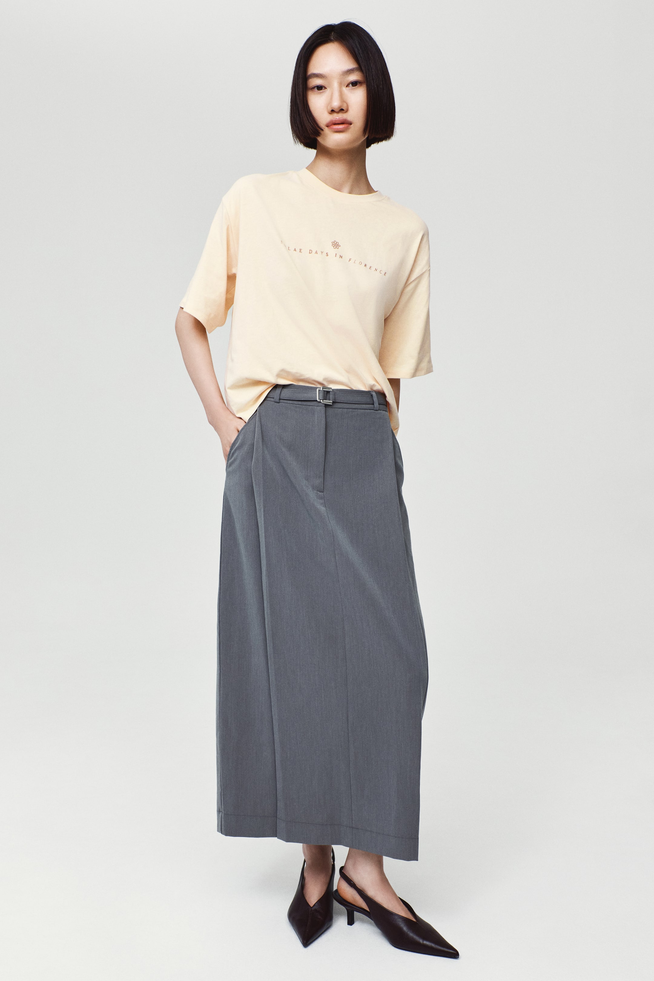 View larger image: A light yellow, oversized t-shirt with dropped shoulders, a round neckline, and a small printed design on the chest is worn by the model. The t-shirt is tucked into a long gray skirt with a belt.