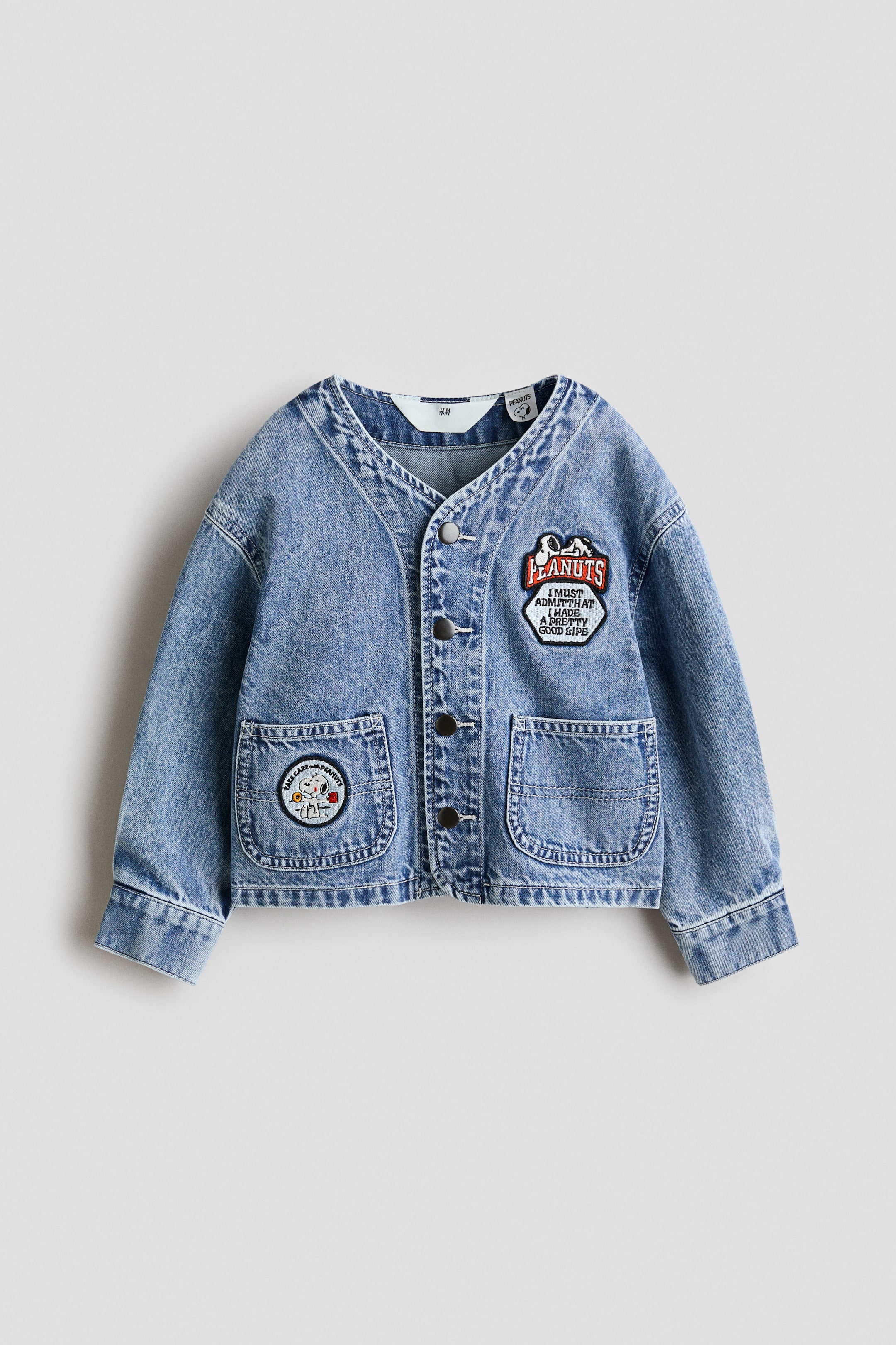 拡大画像を表示: ライトブルーのウォッシュドデニムジャケットが平置きで前面を向くように置かれています。ノーカラーのVネック、前立てはボタン留めでダークカラーのボタンが5つ、裾付近に丸みのある大きなパッチポケットが2つ付いています。右胸には六角形の "Peanuts" ワッペン、左ポケットには丸いSnoopyのベースボールワッペンがあしらわれています。