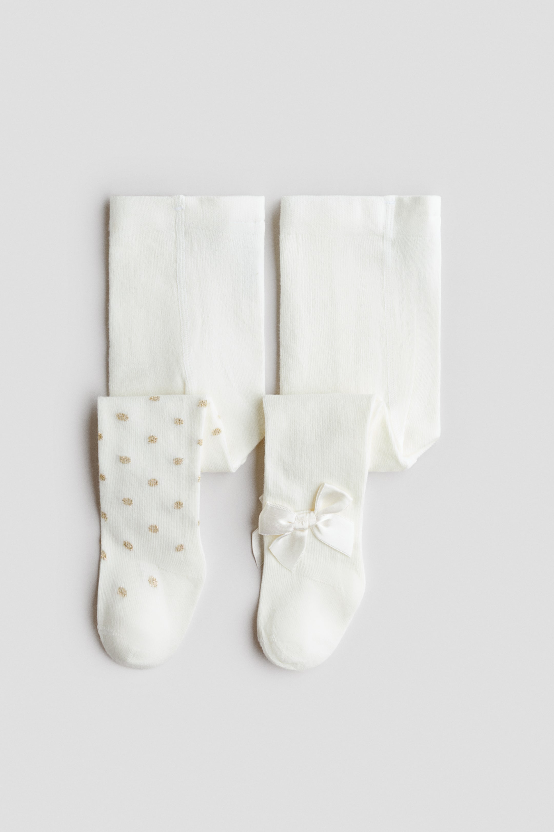 Vis større bilde: To par strikkede strømpebukser i off-white er vist forfra i en flat lay. Det ene paret har gullfargede prikker over hele, mens det andre har et ensfarget design med en hvit satengsløyfe ved ankelen.
