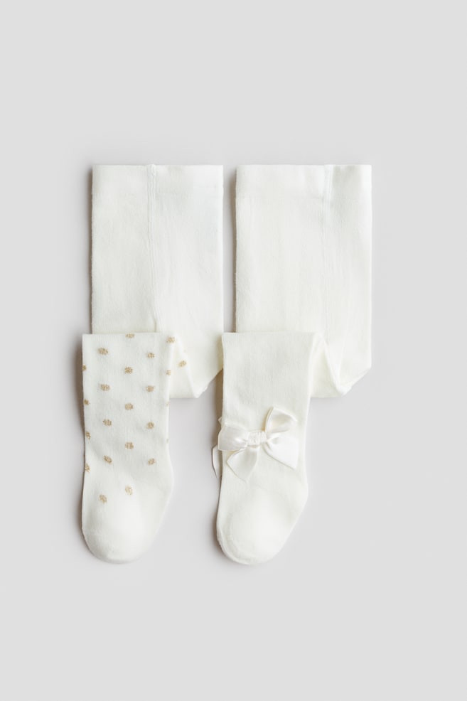 Two pairs of off-white, fine-knit tights are displayed from the front in a flat lay. One pair features all-over gold polka dots, while the other showcases a solid-coloured design accented with a white satin bow at the ankle. - 1