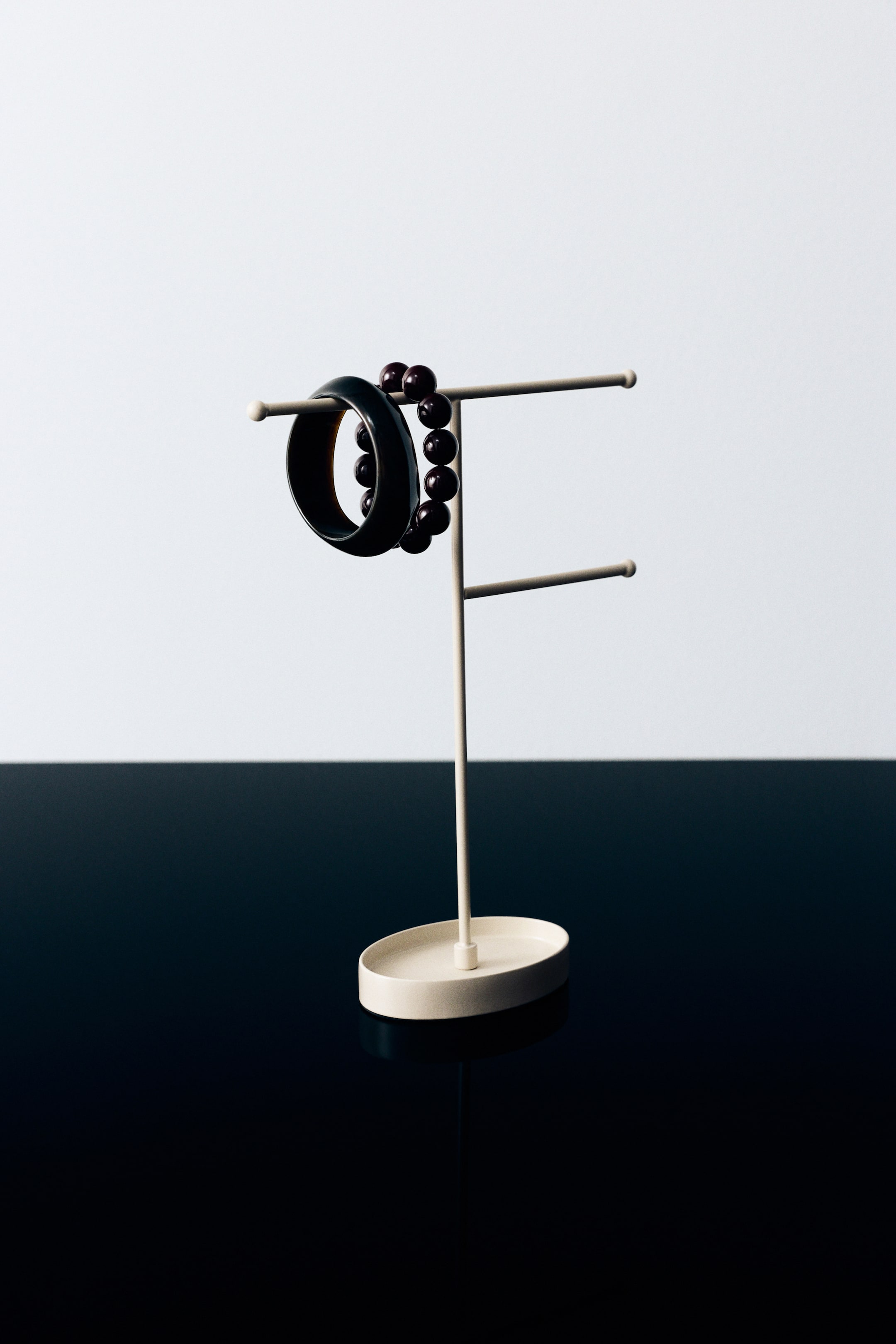 View larger image: A cream-coloured, powder-coated metal jewellery stand is standing upright, featuring a small oval tray base and two horizontal hanging bars extending from a central vertical rod. A thick black bangle and a dark beaded bracelet are hanging on the left side of the top bar.