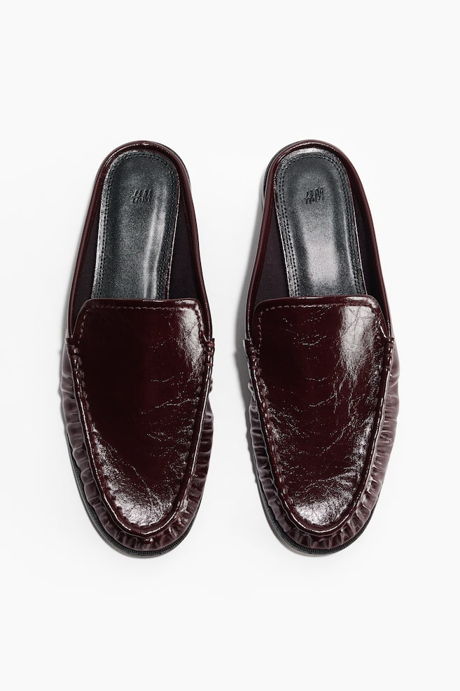 A pair of dark burgundy patent leather loafer mules is displaying a high-shine, crinkled texture on the vamp, with moccasin-style stitching around the rounded toe, presented side-by-side from a top-down view. - 1