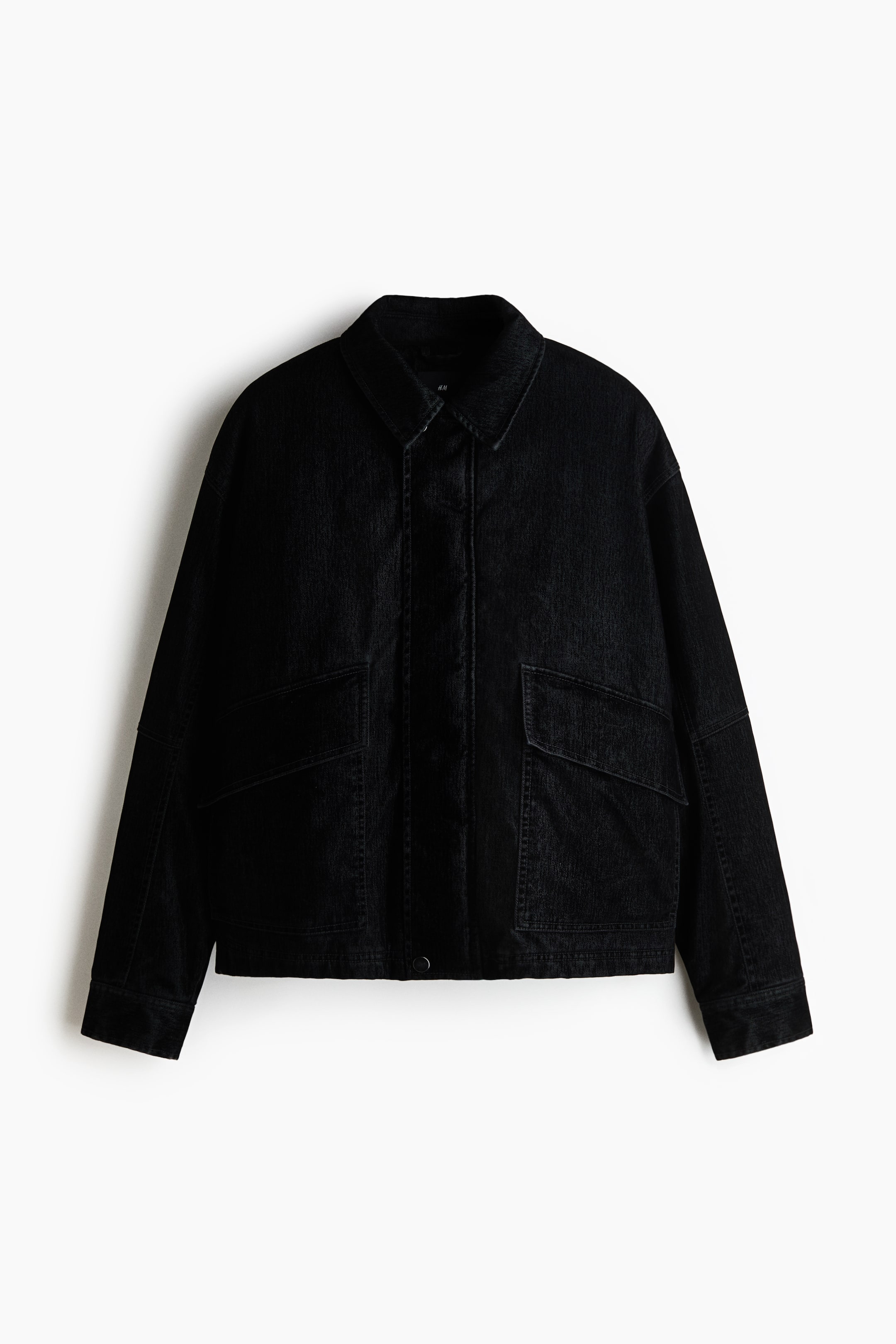 Agrandir l'image: Une veste matelassée noire en sergé de coton est présentée de face, avec un col pointu, des épaules tombantes et une fermeture dissimulée sous un rabat coupe-vent. La veste présente de grandes poches avant avec des rabats en biais et des manches longues avec des poignets simples.
