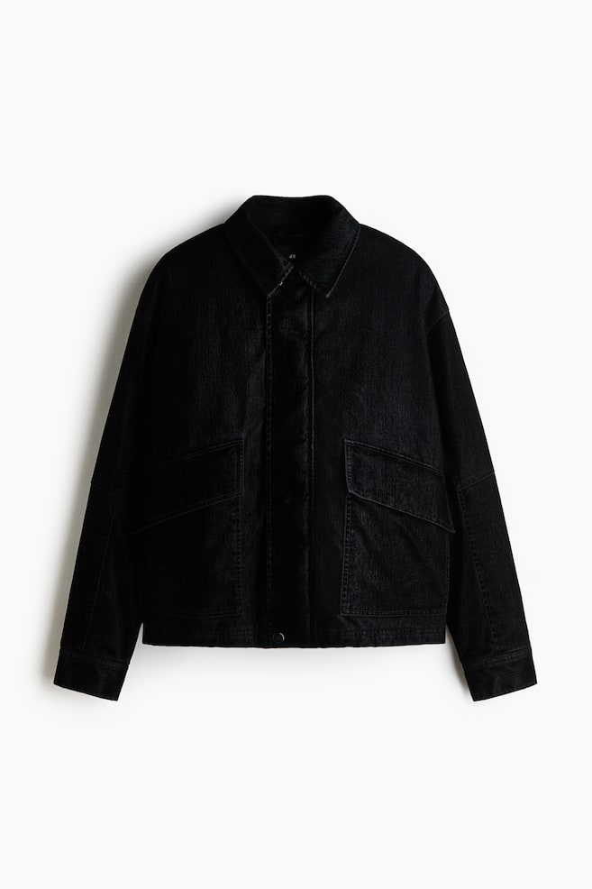 A black padded jacket in cotton twill is shown from a front-facing view, featuring a pointed collar, dropped shoulders, and a concealed front closure under a wind flap. The jacket is displaying large front pockets with angled flaps and long sleeves with simple cuffs. - 1