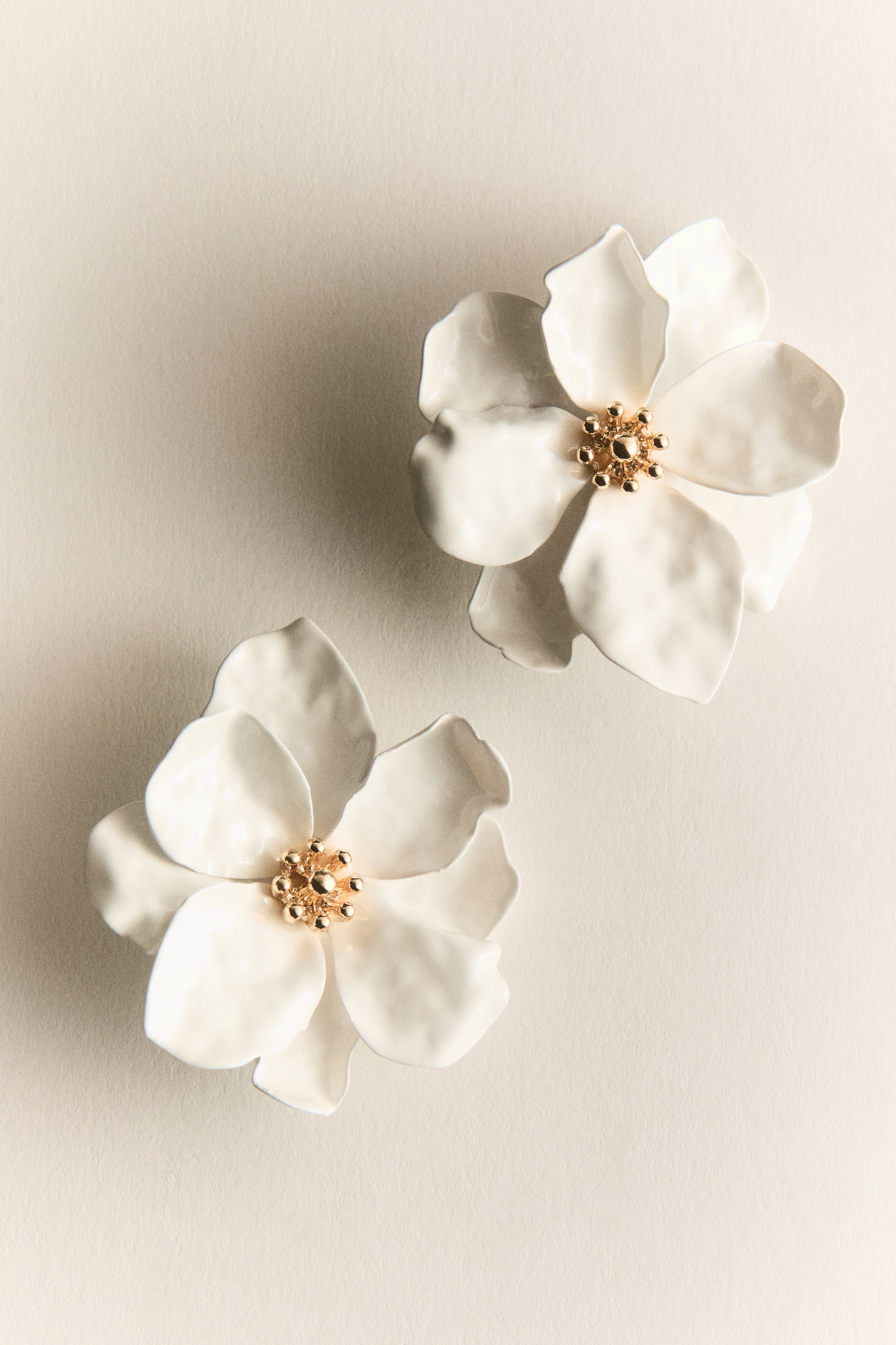 Agrandir l'image: Deux boucles d'oreilles en forme de fleur sont posées à plat, chacune présentant plusieurs pétales blancs sculptés, légèrement incurvés et irréguliers, entourant un centre perlé doré.