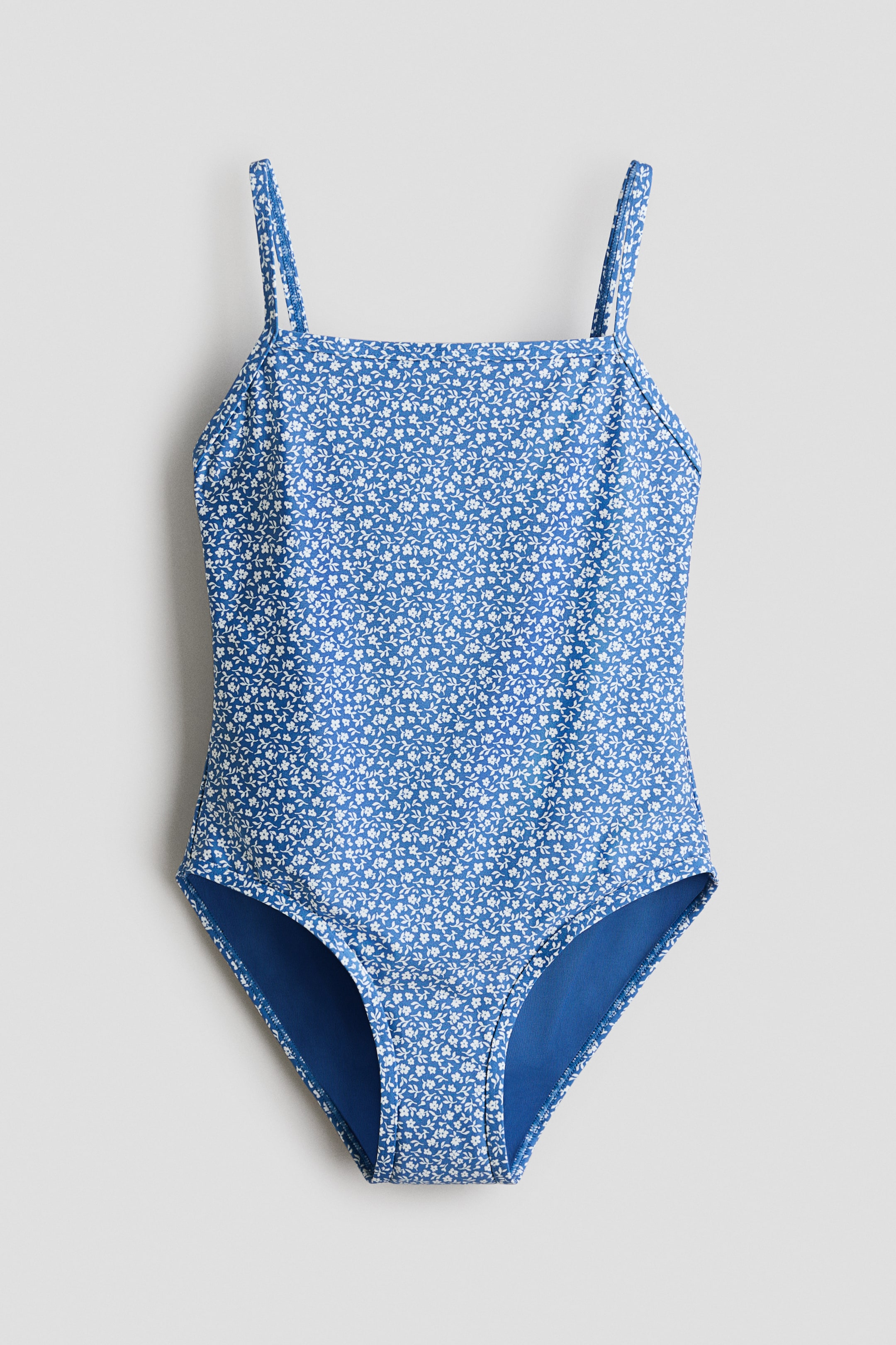 View larger image: One-piece swimsuit shown from the front, featuring a blue base with a dense white floral pattern. It is designed with thin spaghetti straps and a square neckline. The leg openings are trimmed with solid blue fabric.