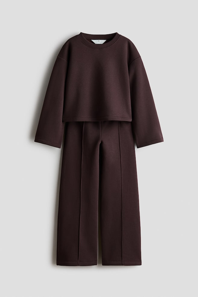 A dark brown fleece sweater and track pants set is shown in a front-facing view. The cropped sweater is featuring a ribbed crew neck, dropped shoulders, and wide sleeves, while the matching wide-leg pants are displaying prominent sewn-in creases running down the front of each leg. - 1