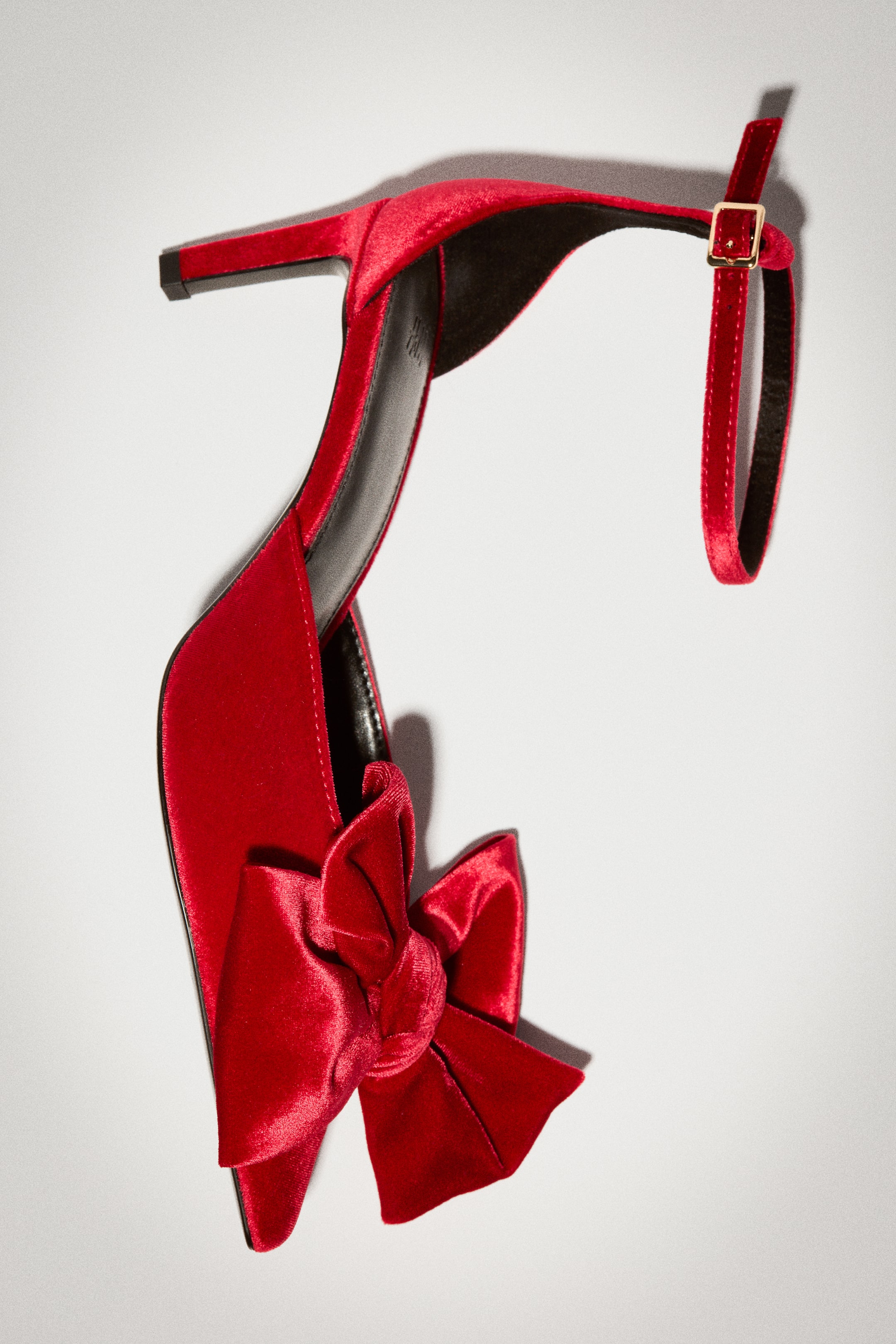 View larger image: A red velour high-heeled shoe is shown from a top-down angle, featuring a pointed toe adorned with a large, decorative bow. The design includes a narrow, covered heel, a smooth black lining, and an adjustable ankle strap with a gold-tone buckle.