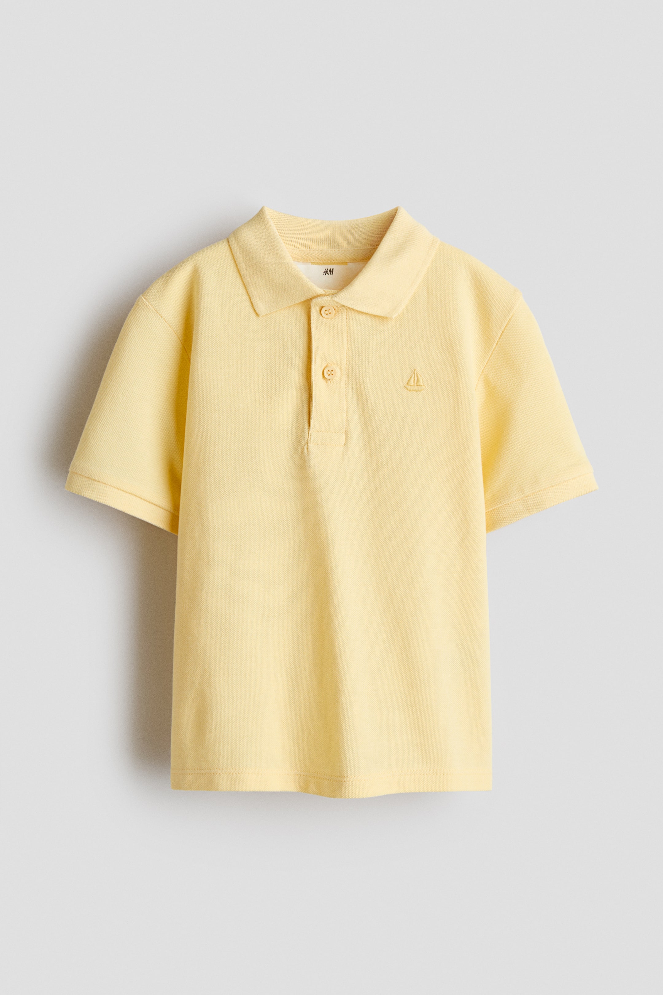 View larger image: A light-yellow polo shirt is shown from the front, featuring a ribbed collar, a two-button placket and short sleeves with ribbed cuffs, and a small embroidered sailboat emblem on the left chest.