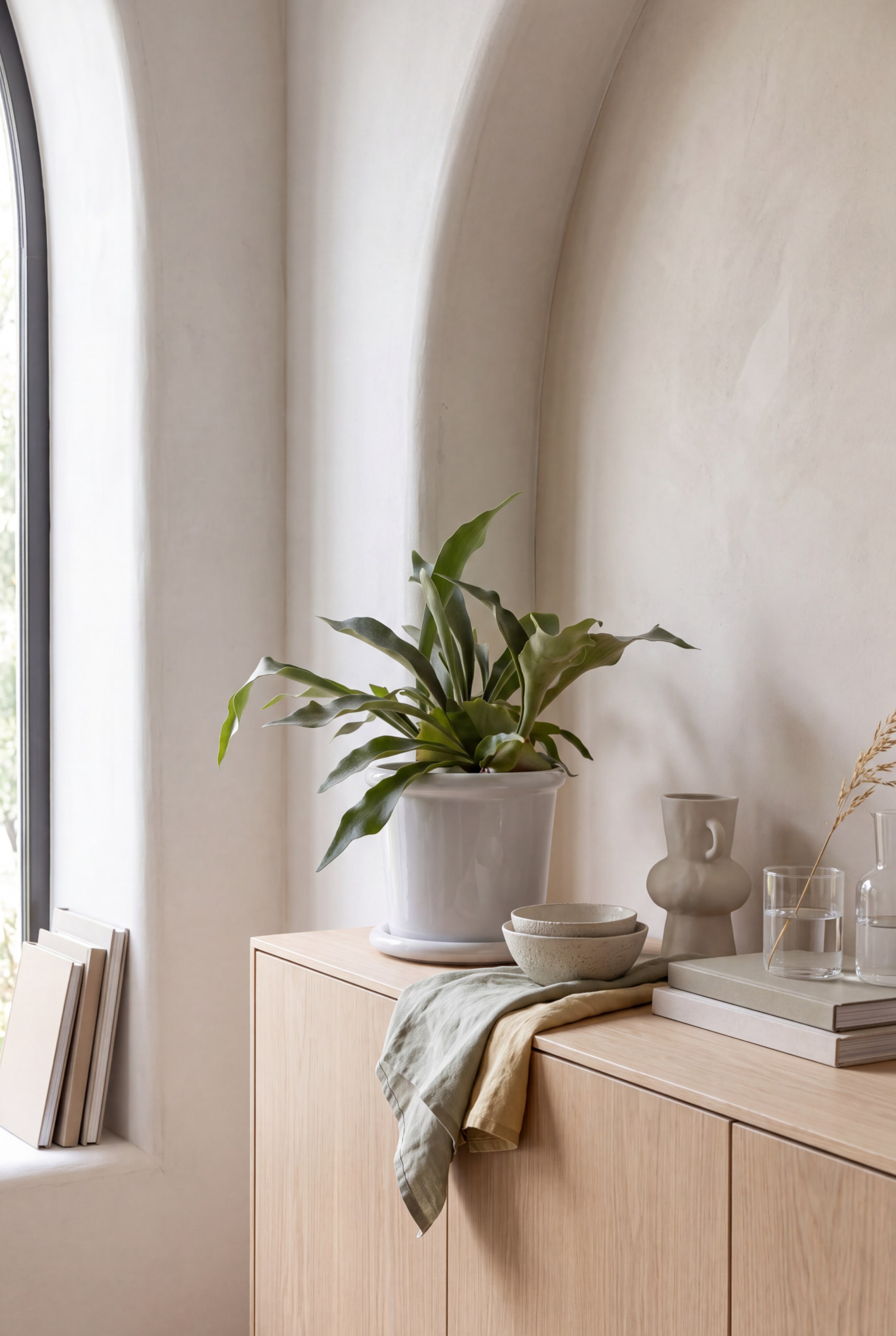 Visualizza immagine più grande: Un vaso in ceramica smaltata bianca con sottovaso coordinato contiene una pianta verde a foglia con lunghe fronde arcuate. Il vaso è appoggiato su un mobile in legno chiaro, accanto a ciotole in ceramica impilate e tessuti di lino drappeggiati.
