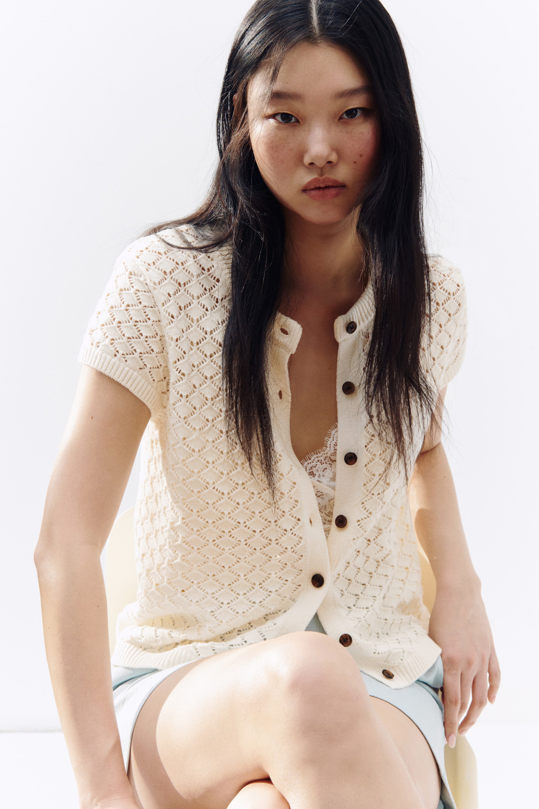 Prikaži veću sliku: Kremasti kardigan sa kratkim rukavima nosi se, sa otvorenim pletenim dijamantskim uzorkom. Ima prednji deo na kopčanje sa okruglim braon dugmićima i rebrastim manžetnama i porubom, složen preko belog čipkanog braleta.