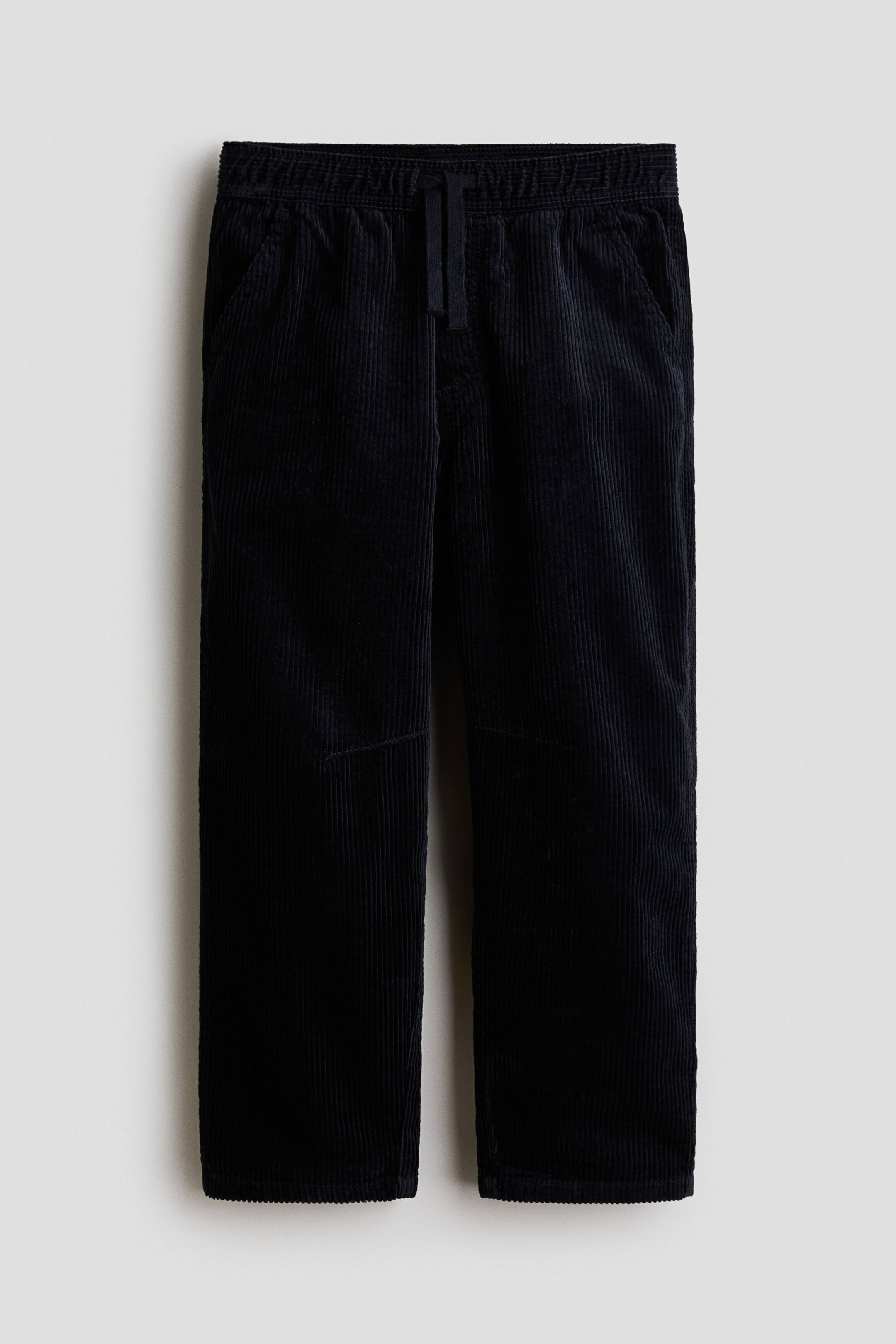 Grotere afbeelding bekijken: Een zwarte corduroy jogger wordt van voren getoond, met een gestructureerde geribbelde stof, een elastische tailleband met verstelbaar trekkoord en schuine zijzakken. De broek is ontworpen met een nepgulp en een relaxte barrel-leg silhouette.