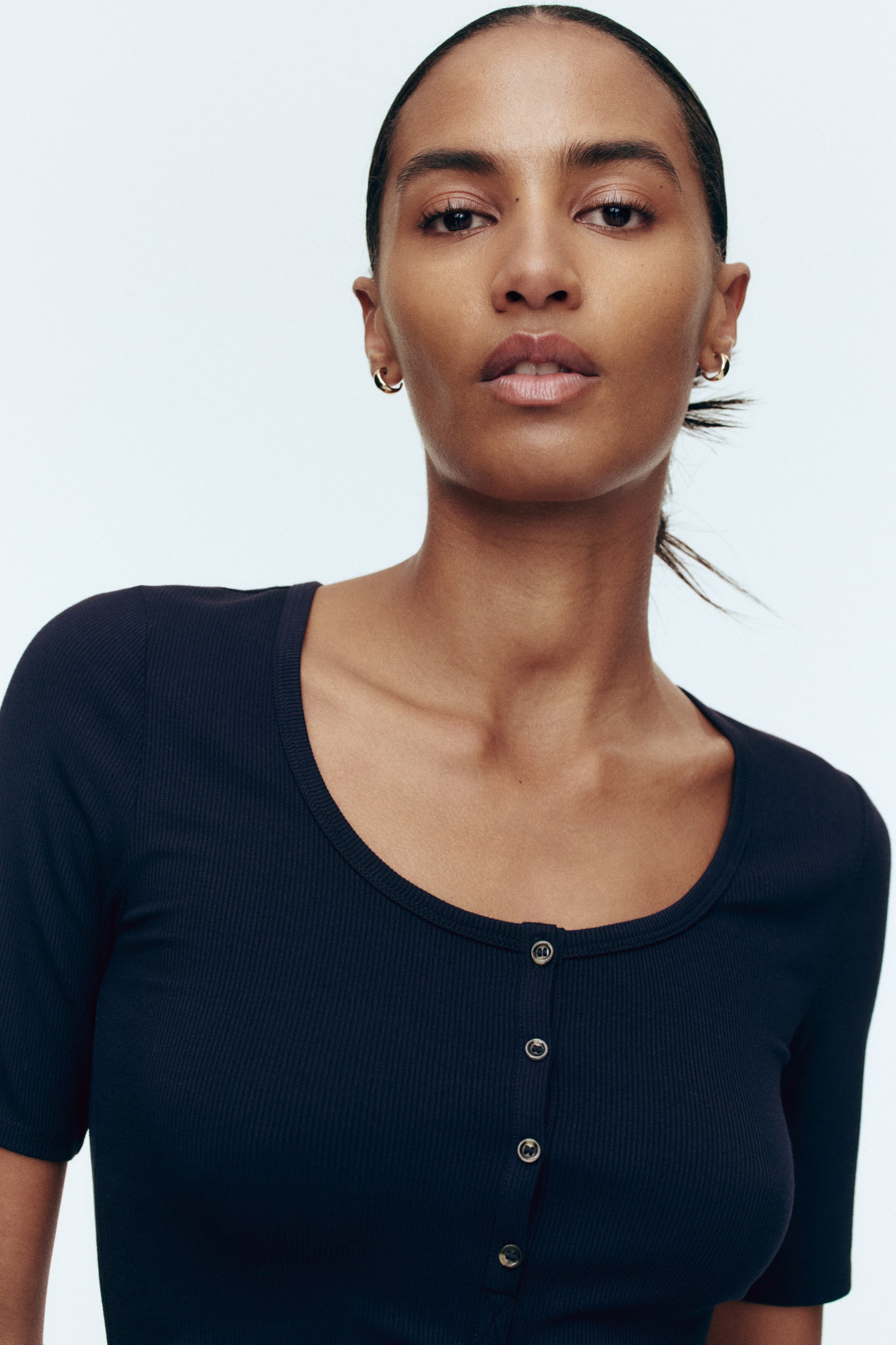 Xem ảnh lớn hơn: Chiếc áo xanh đậm có gân với tay ngắn có điểm nhấn là cổ áo tròn và hàng nút phía trước, được gắn các nút màu bạc.