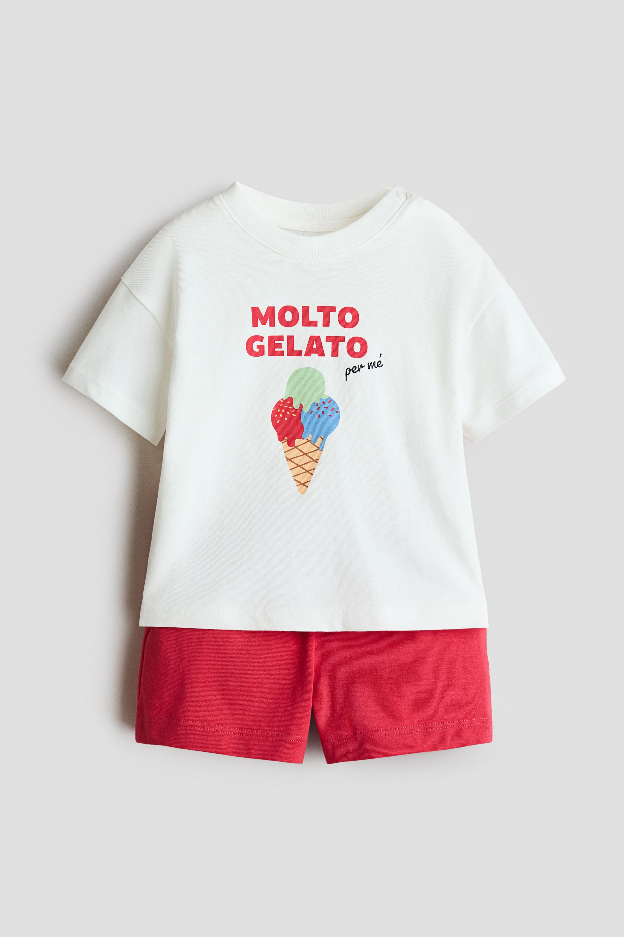 View larger image: This two-piece cotton set features a white, short-sleeved T-shirt and solid red shorts shown from the front. The T-shirt displays a graphic of a three-scoop ice cream cone beneath the red and black text "MOLTO GELATO per me", while the matching red shorts complete the outfit.