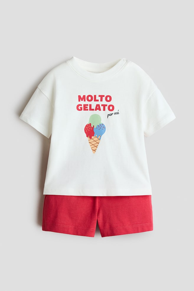 This two-piece cotton set is featuring a white, short-sleeved t-shirt and solid red shorts shown from a front angle. The t-shirt is displaying a graphic of a three-scoop ice cream cone beneath the red and black text "MOLTO GELATO per me", while the matching red shorts are completing the outfit. - 1