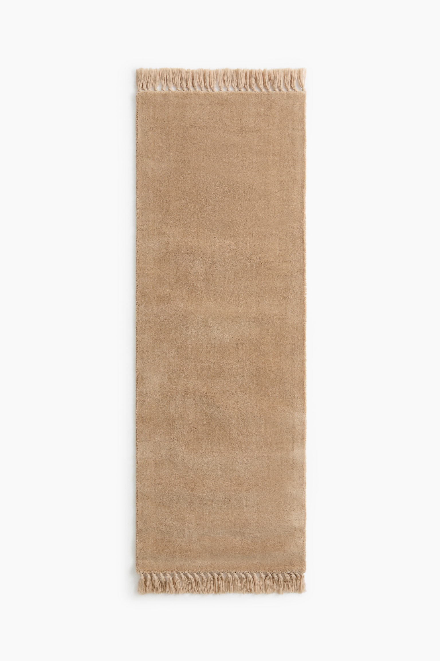A rectangular beige runner rug is presenting in a front-facing view, featuring a soft, plush texture and a solid color. The rug is displaying matching beige fringes along both of its shorter ends.