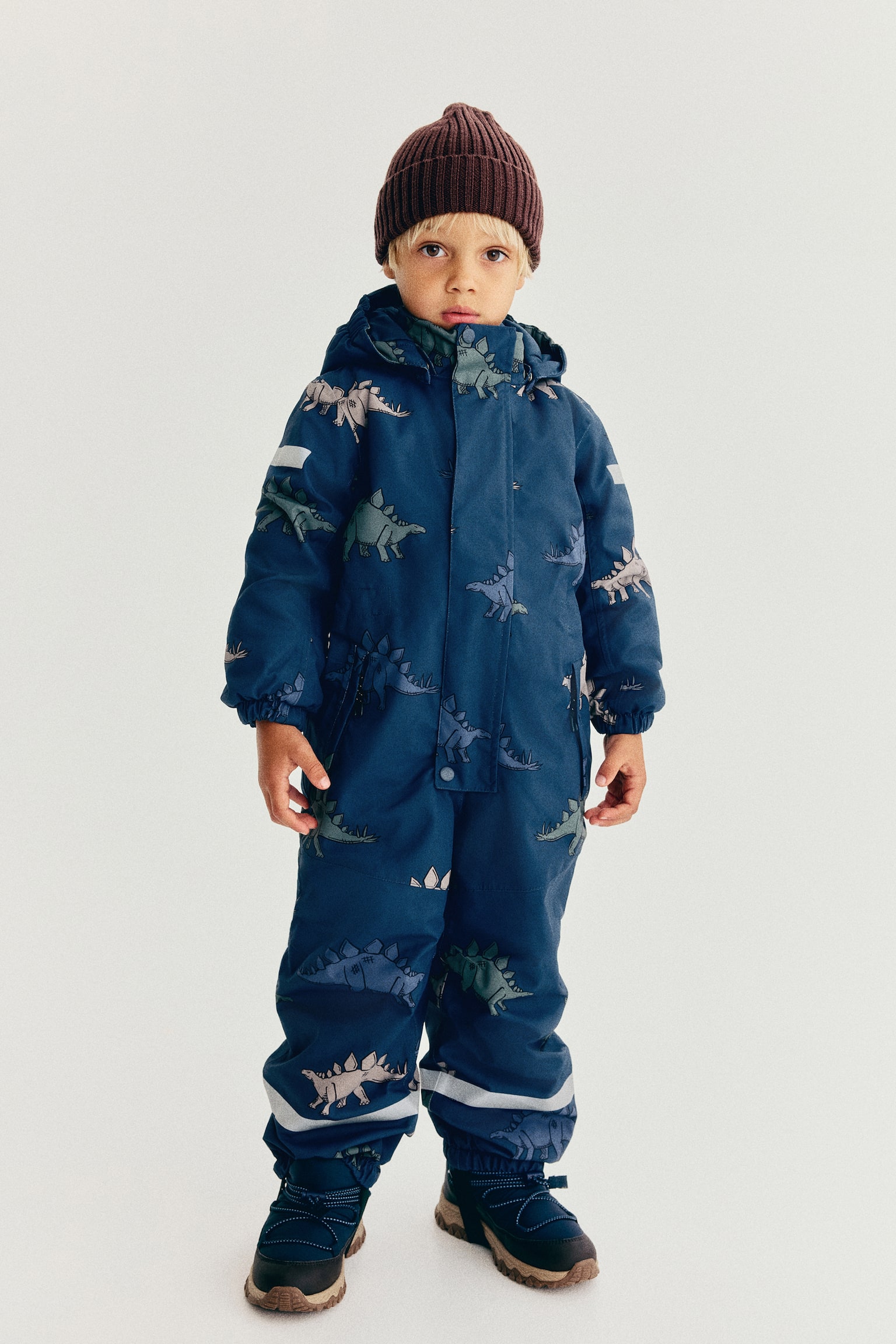 A children's navy blue all-in-one snowsuit is shown front-facing, featuring an all-over print of stegosaurus dinosaurs in muted beige, green and purple. The garment includes an attached hood, a central zip fastening concealed beneath a snap-button storm flap, reflective safety bands on the upper arms and lower legs, and black elastic stirrups extending from the elasticated cuffs. - 1