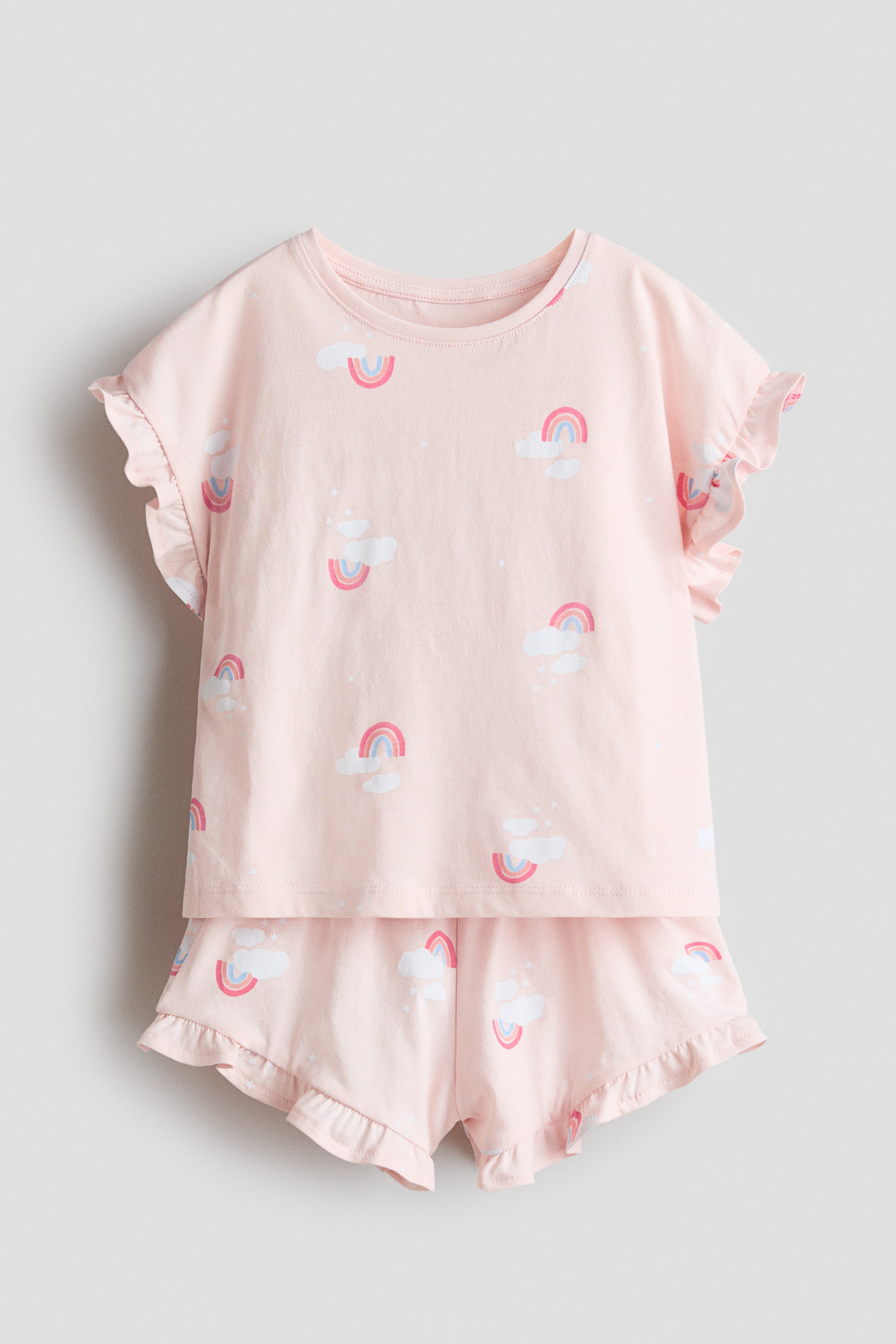 이미지 확대 보기: 핑크색 프린트된 면 저지 파자마 세트가 앞에서 소개됩니다. 이 세트는 둥근 목선과 프릴 장식의 짧은 소매가 있는 상의와, 허리 밴드와 프릴 장식의 밑단이 있는 반바지로 구성되어 있으며, 무지개와 구름 패턴이 돋보입니다.