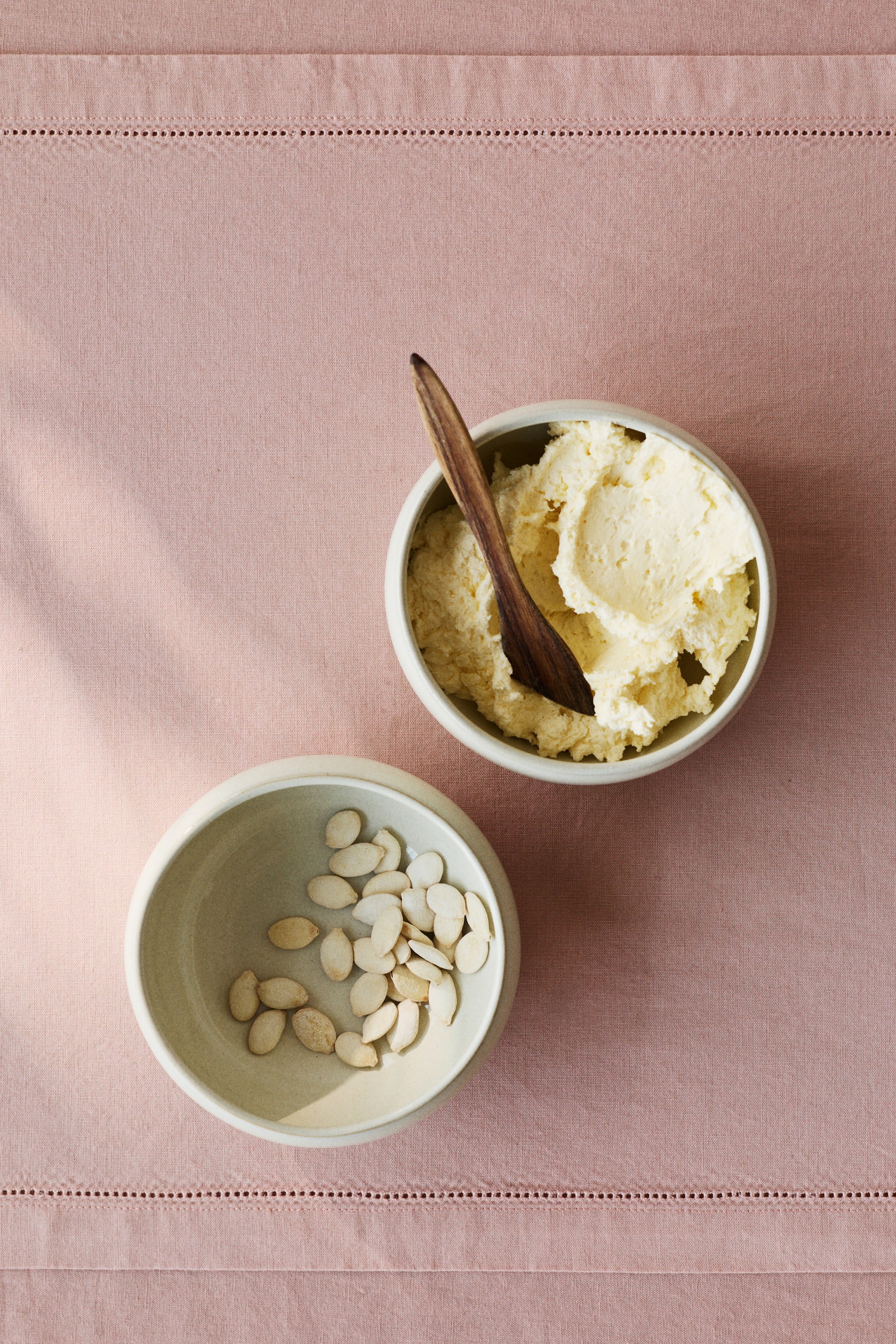 View larger image: Two round, light-coloured ceramic bowls are presented from an overhead view; one is holding a creamy, textured pale yellow substance with a wooden spoon resting inside, and the other contains numerous pale, oval-shaped seeds.