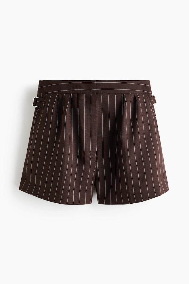 Los pantalones cortos de color marrón oscuro presentan finas rayas verticales blancas y una cintura lisa. Hebillas ajustables de tono plateado son visibles a cada lado de la cintura, y pliegues se forman debajo de la cintura, otorgando a los pantalones cortos una estructura entallada. - 1