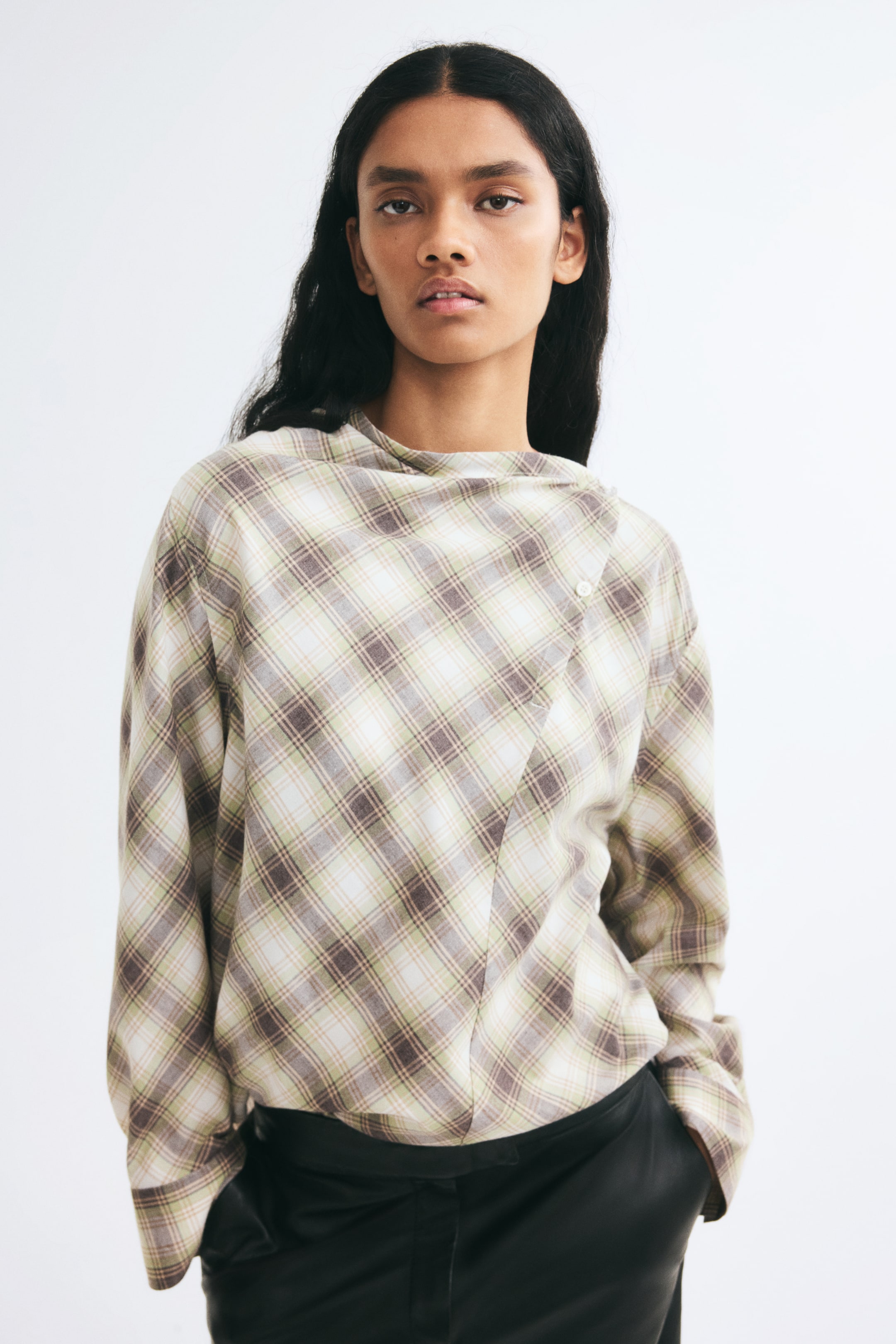 Agrandir l'image: Une blouse tissée à carreaux dans des tons de vert, de marron et de crème est portée par le mannequin, rentrée dans un pantalon noir. La blouse présente un col bénitier montant, des manches longues et un devant portefeuille asymétrique fermé par un bouton près de l’épaule.