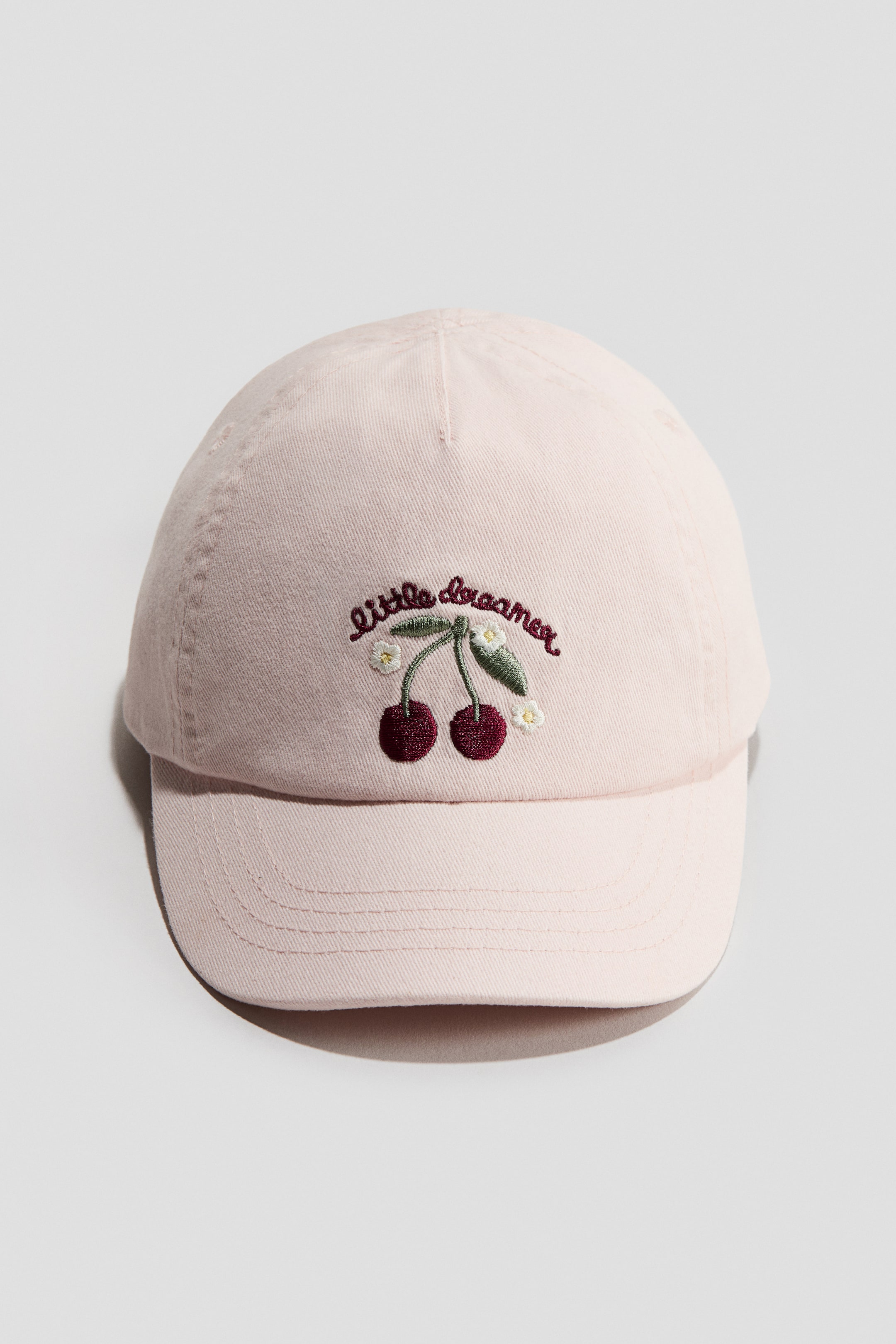 View larger image: A light pink cap in a cotton weave is facing FRONT, featuring an embroidered design on the front with the text 'little dreamer' above two cherries, leaves, and small white flowers.