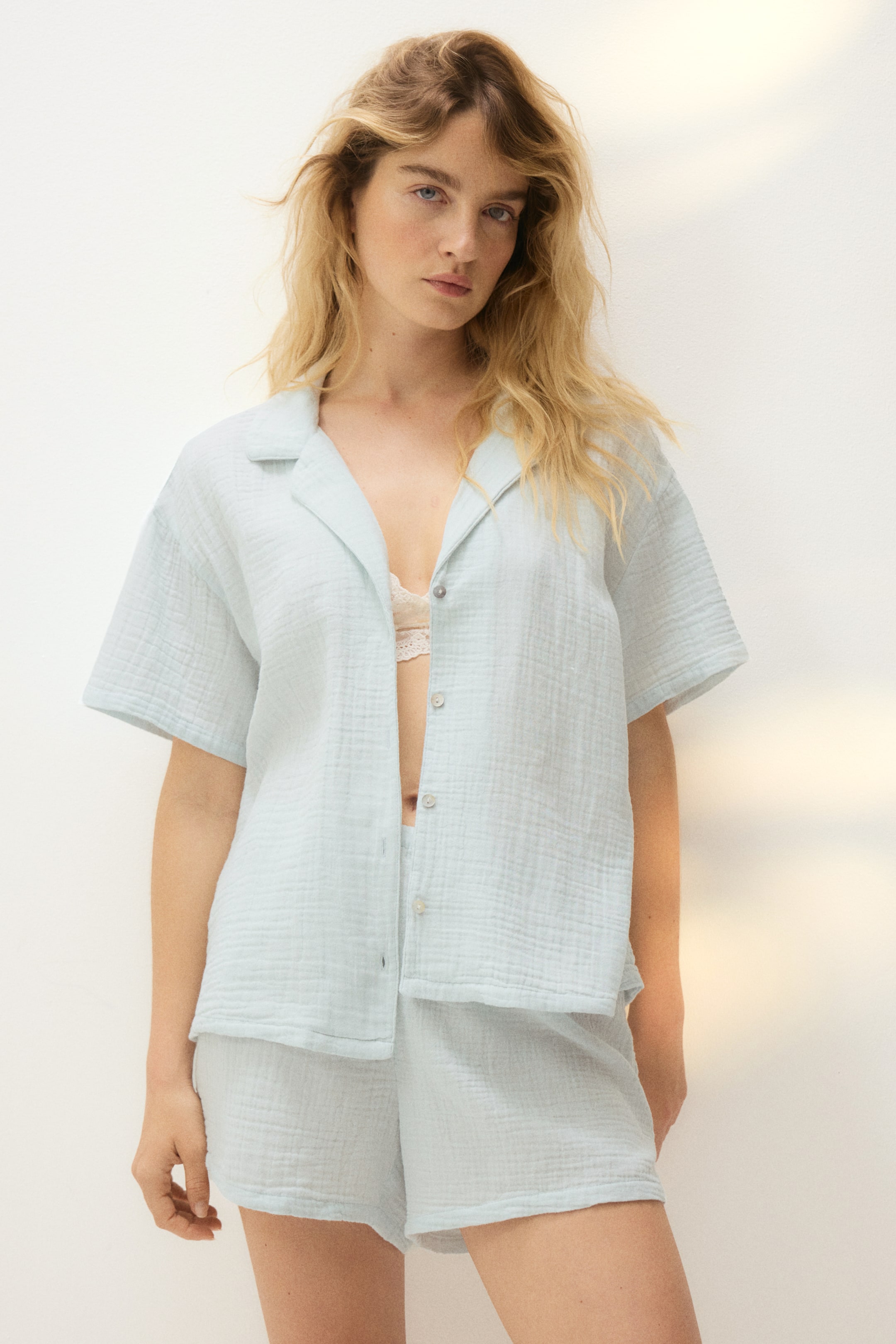 View larger image: A light blue crinkled fabric short-sleeved shirt paired with matching shorts is showcased. The shirt features an open collar design with visible buttons, several of which are undone to reveal a lace bralette underneath.