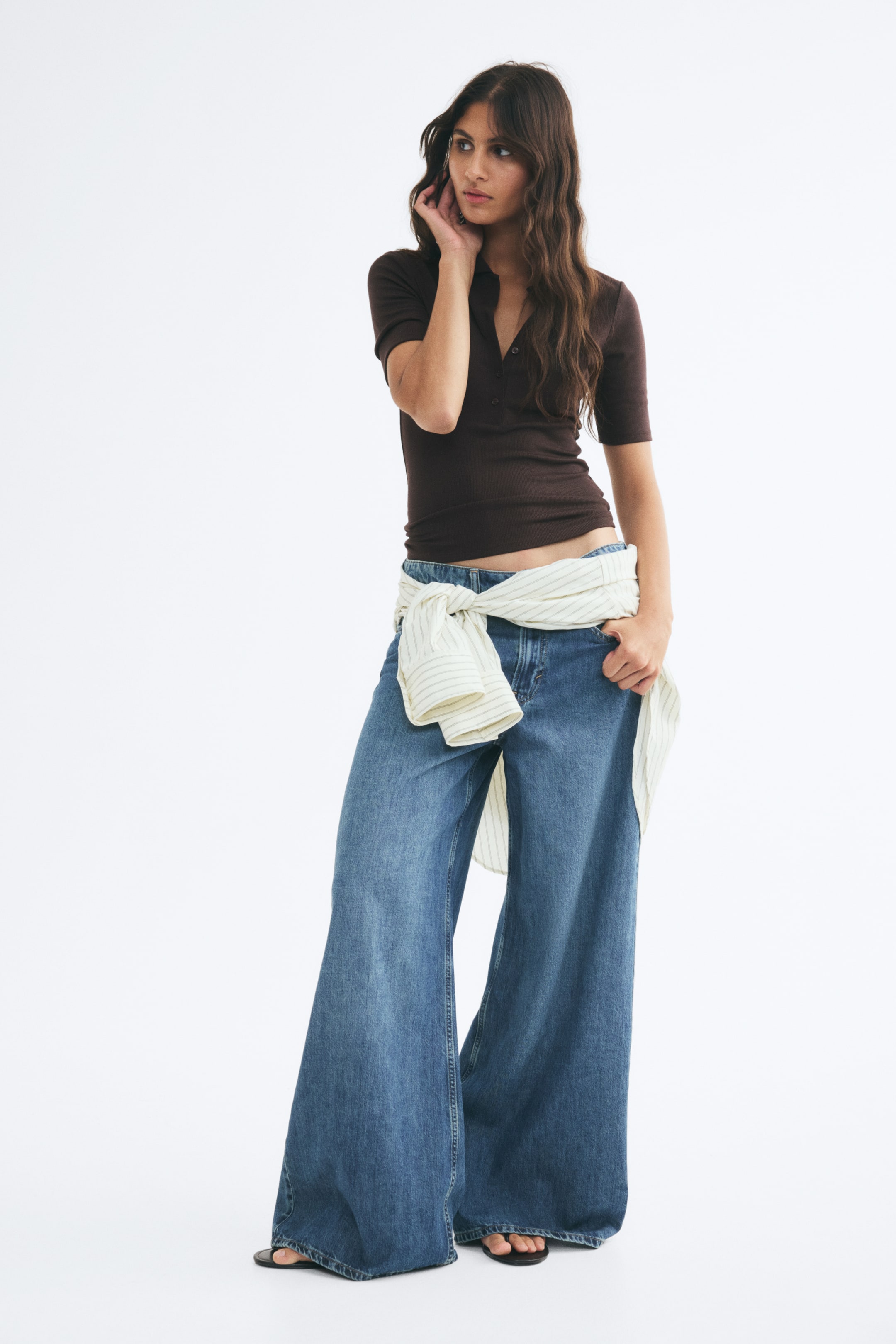 View larger image: A dark brown fitted polo shirt in a soft rib knit is worn by the model. The short-sleeved shirt features a collar and button placket, and is styled with wide-leg blue jeans and a striped shirt tied around the waist.