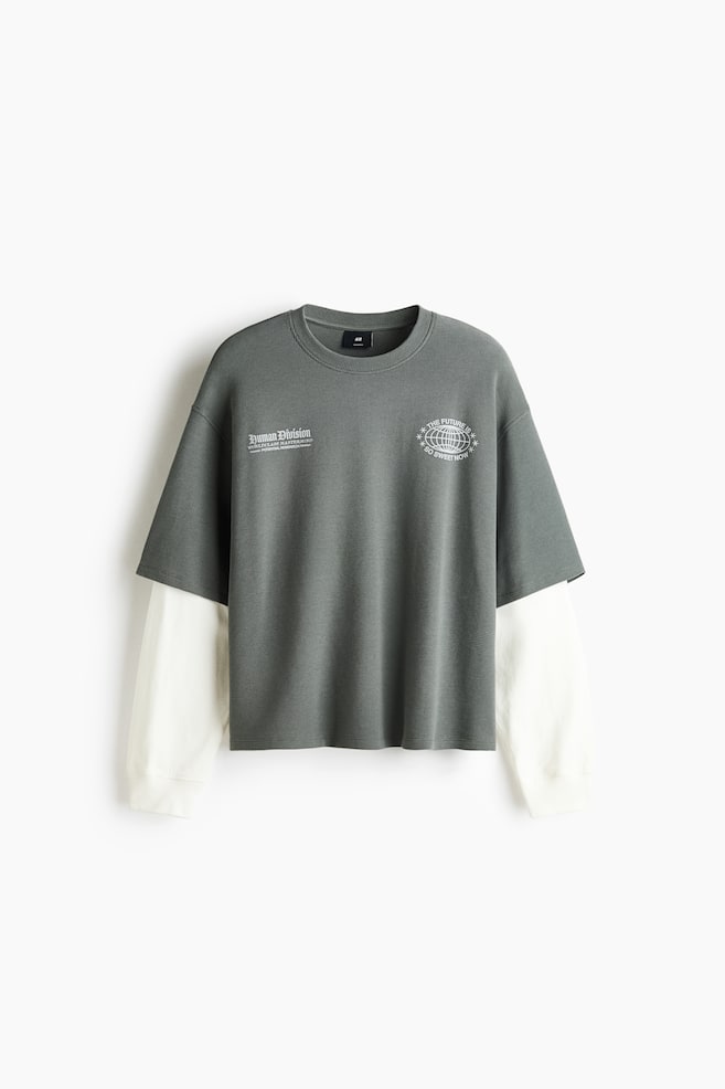 Uma t-shirt de manga comprida em camadas estÃ¡ voltada para a frente, exibindo uma camada exterior de manga curta em cinzento-escuro com textura, com mangas compridas em creme a contrastar, a sair de baixo. A camada exterior apresenta duas estampas grÃ¡ficas brancas no peito e gola redonda canelada, enquanto as mangas em creme terminam em punhos canelados. - 1