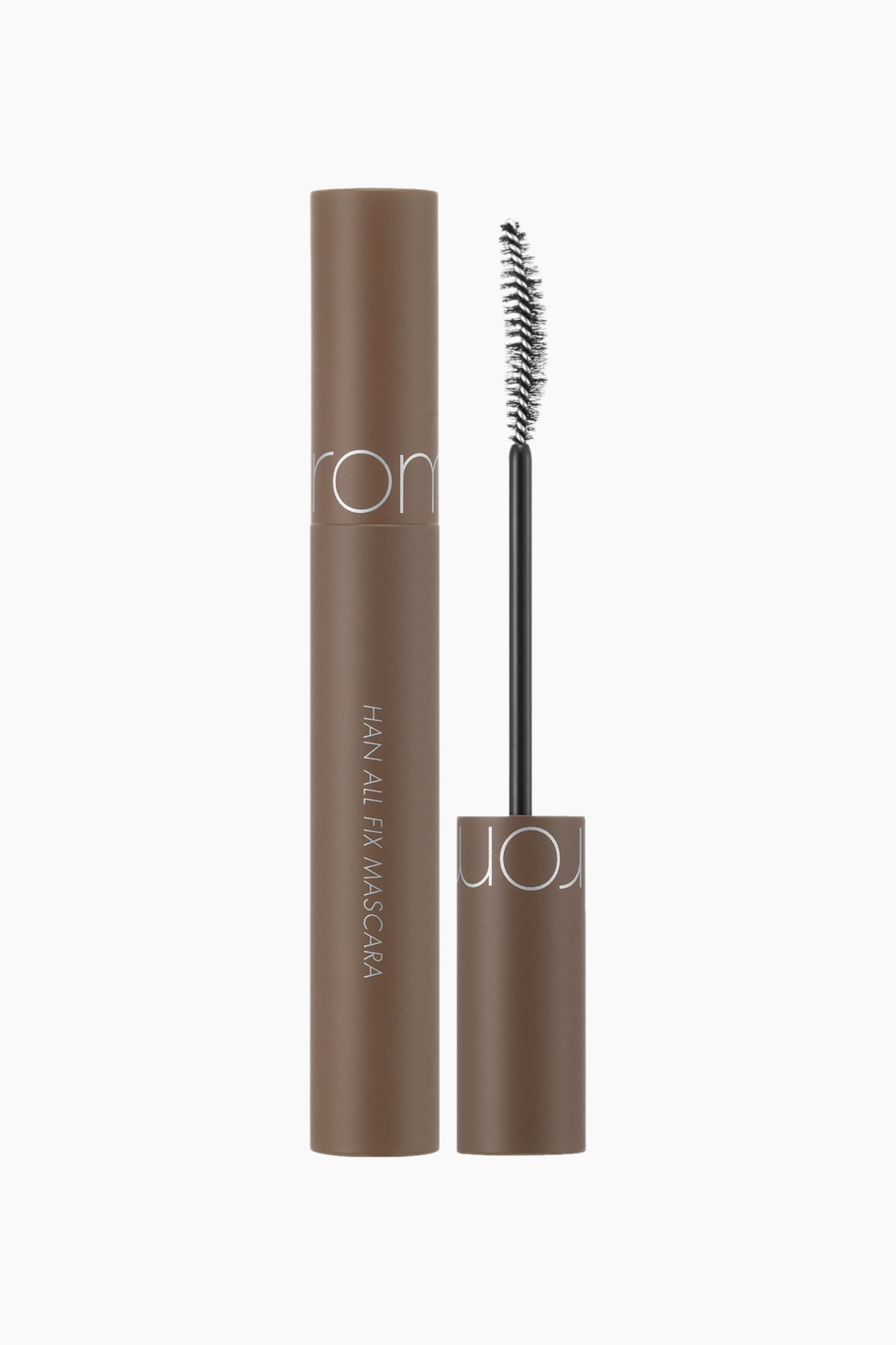 Vis større bilde: En brun, sylindrisk mascara-tube står oppreist, med «rom&nd» og «HAN ALL FIX MASCARA» synlig i sølvfarget skrift. Lokket er tatt av og står ved siden av, og viser en svart mascarabørste med en buet, bustete børste.