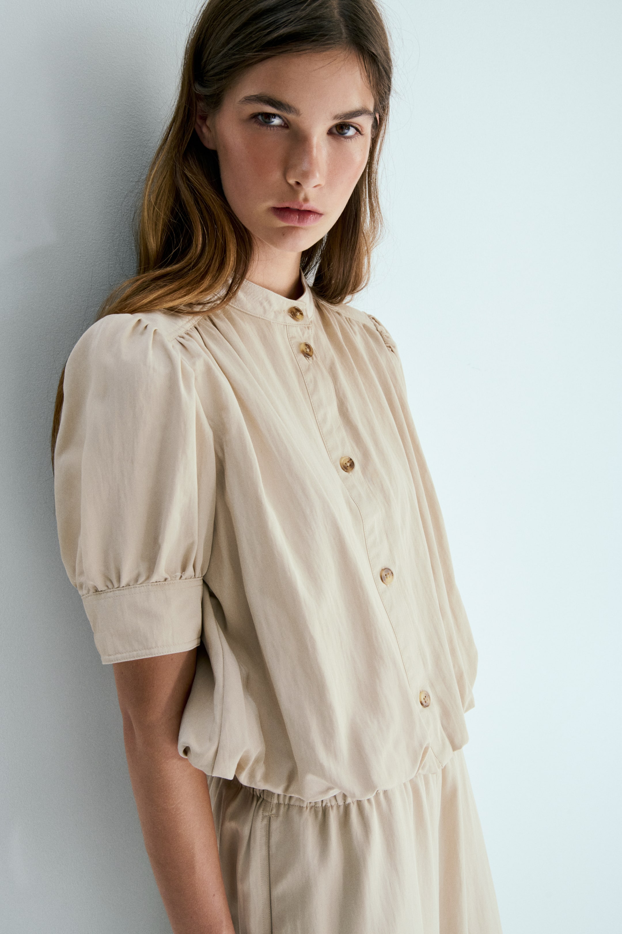 View larger image: A light beige top paired with matching bottoms is displayed. The top boasts a band collar, a front button placket adorned with brown buttons, and puffed sleeves with wide cuffs. Its elasticized hem creates a blouson effect over the elasticized waistband of the matching bottoms.